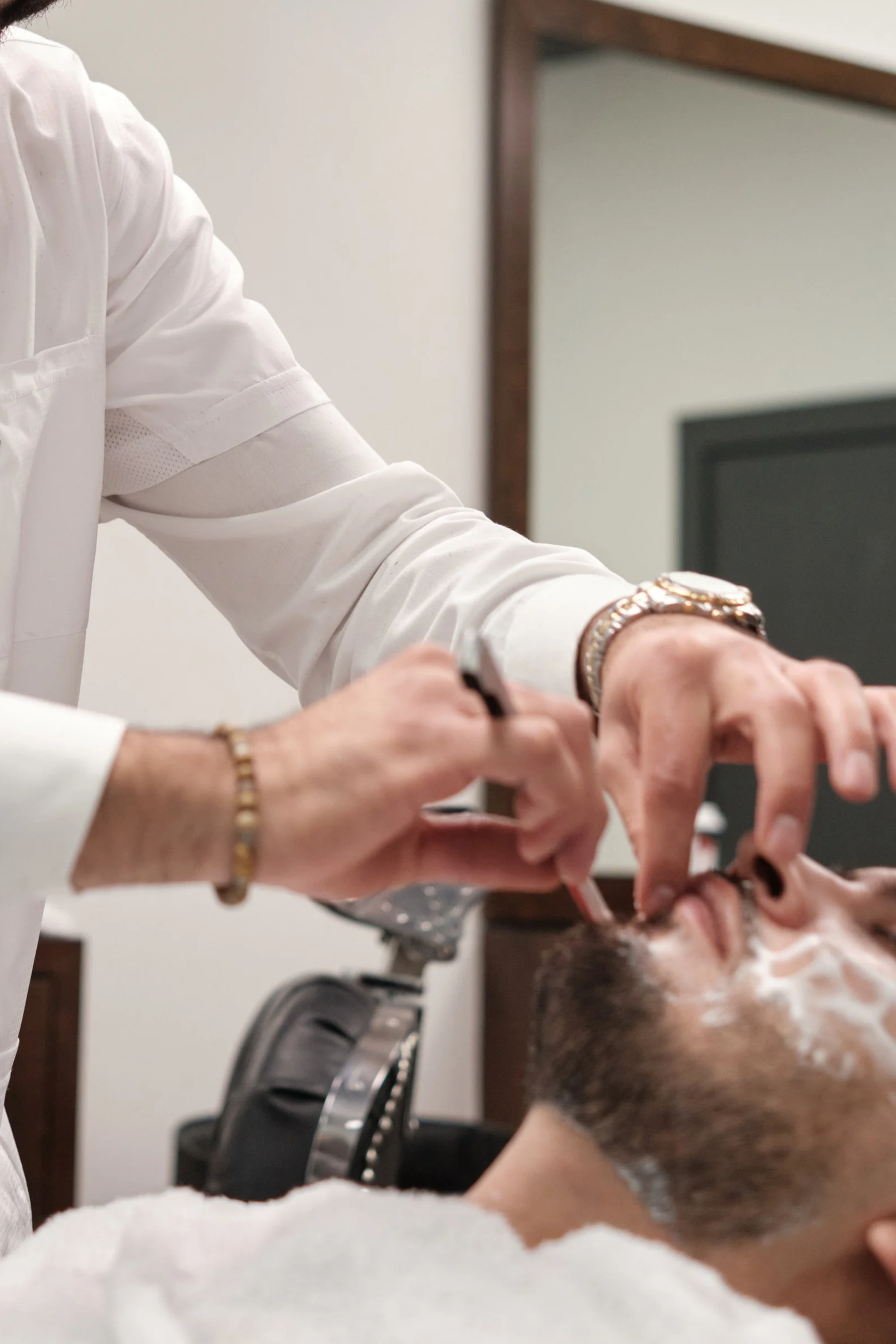 A person receiving a shave at a barbershop, lying back with a white towel around their shoulders while a barber applies shaving cream to their face.