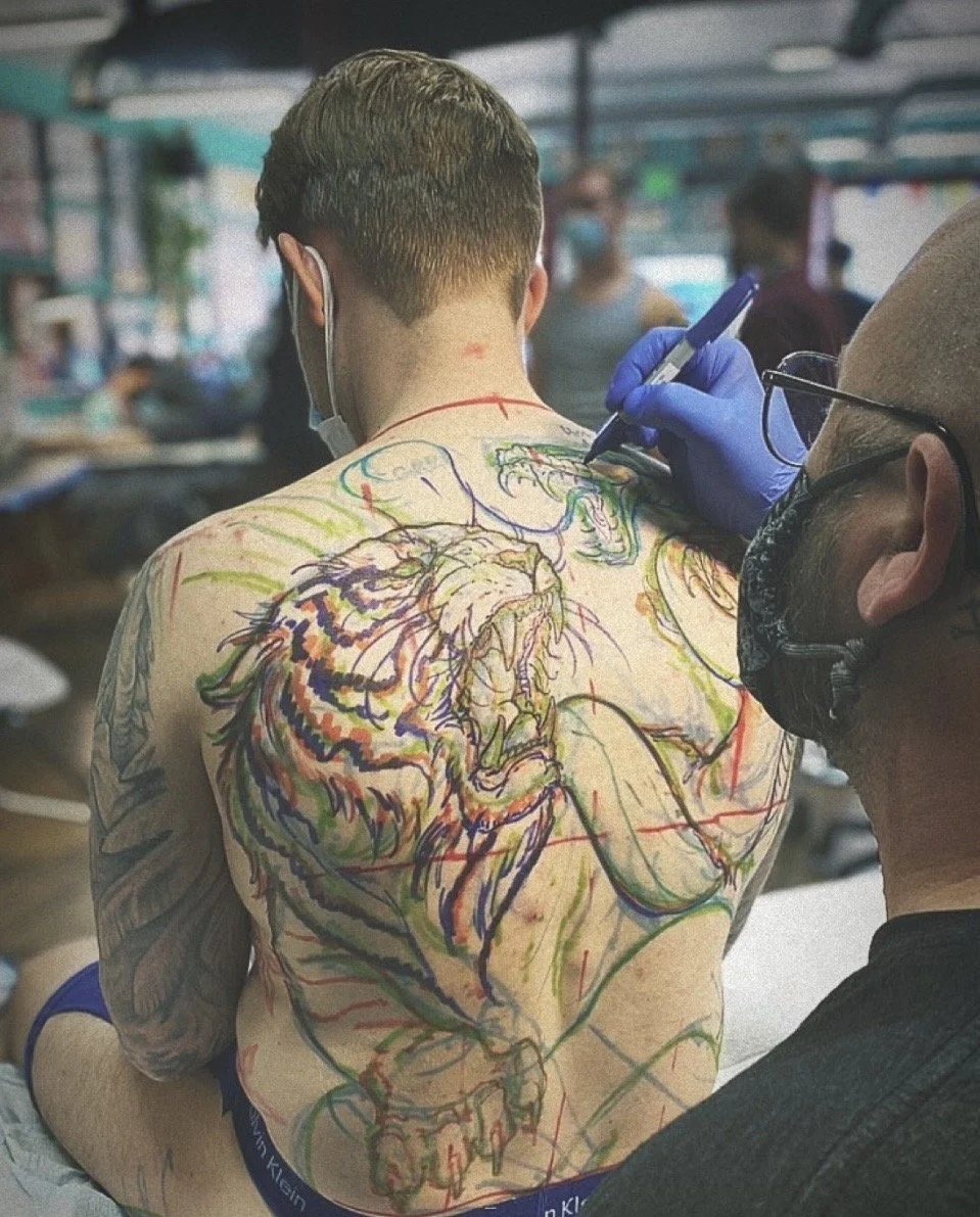 A person with a large, colorful tattoo of a roaring tiger on their back inside a tattoo parlor, receiving a tattoo from an artist wearing glasses and a mask, with other people and artwork visible in the background.