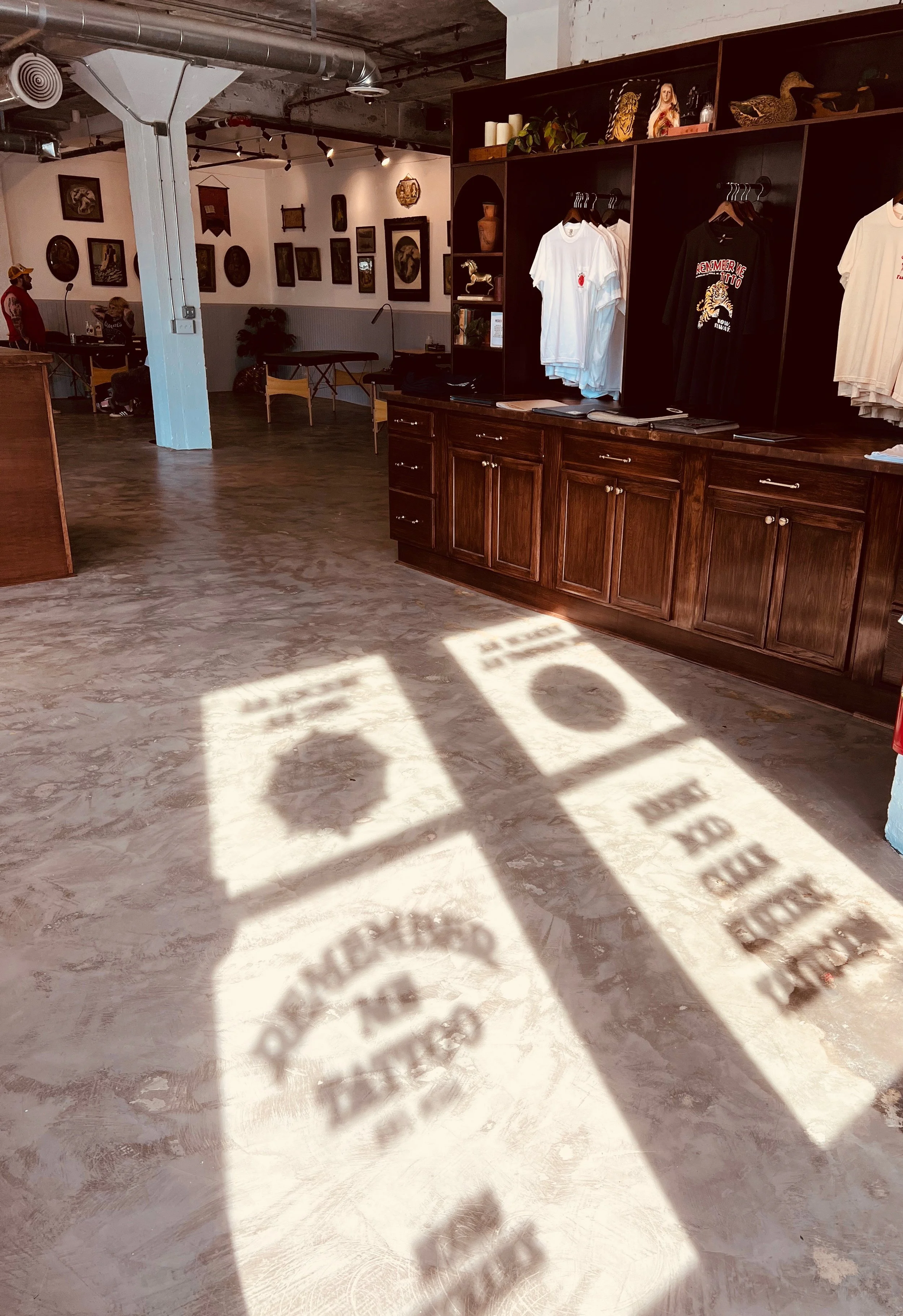 Interior of a retail space with wooden cabinets, clothing on hangers, and shadow patterns on the polished concrete floor created by sunlight through windows.