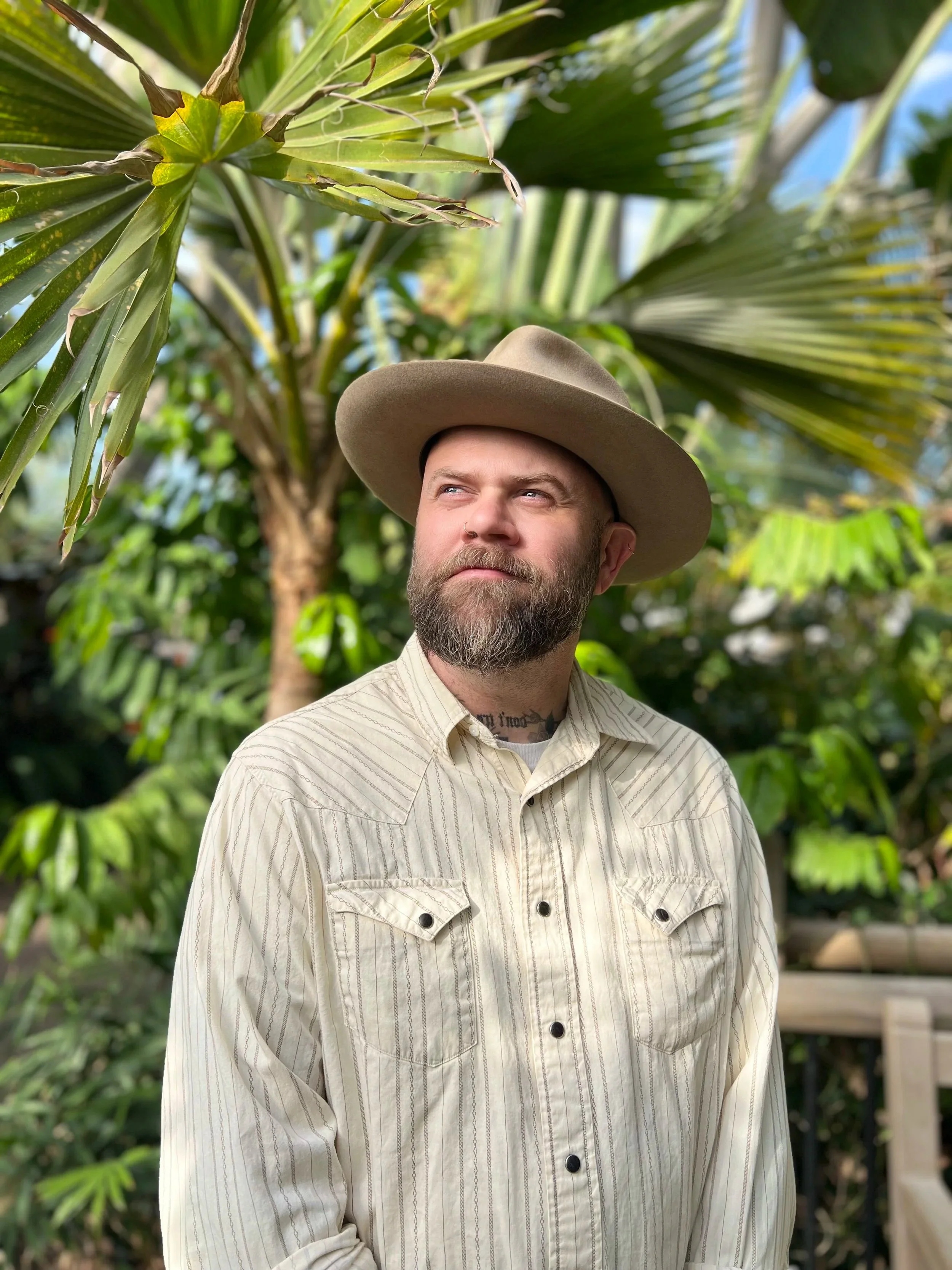 A man with a beard wearing a beige, striped button-up shirt and a wide-brimmed beige hat stands outdoors among lush green tropical plants, with a slight smirk and looking off to the side.