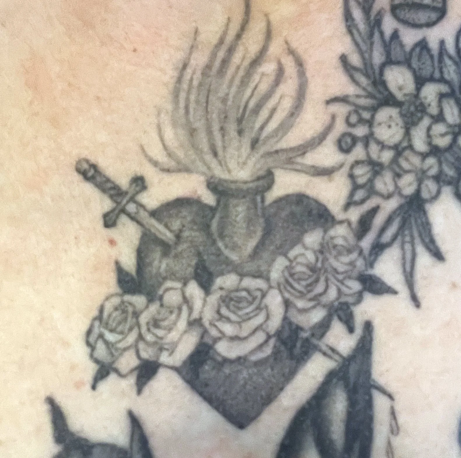 Black and white tattoo of a flaming torch, roses, a heart, a sword, and flowers.