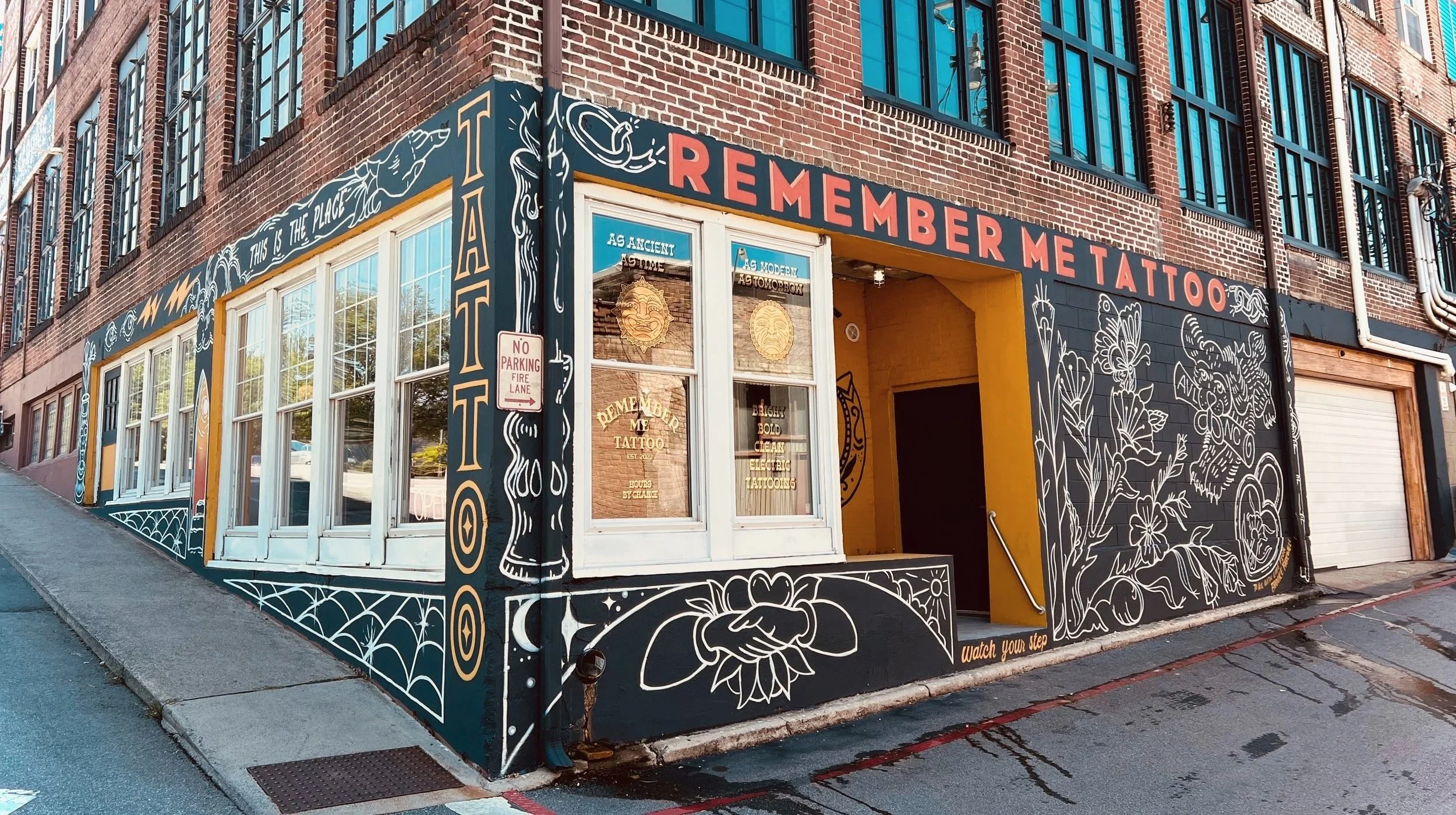 Street corner tattoo shop with colorful mural art on the exterior walls depicting flowers, animals, and decorative elements; sign reads 'Remember Me Tattoo'.