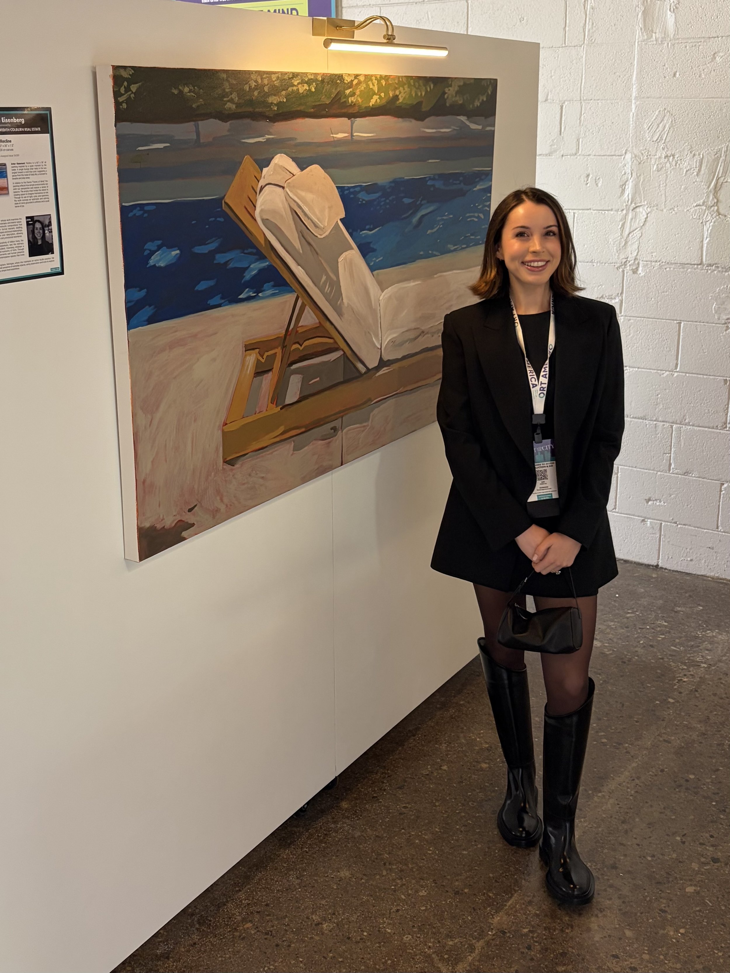 A young woman with shoulder-length brown hair, wearing a black blazer, long skirt, and tall black boots, standing in front of a painting at an art gallery, smiling at the camera.