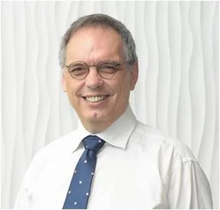A smiling man with glasses, wearing a white shirt and navy blue tie with white polka dots, standing in front of a textured white background.