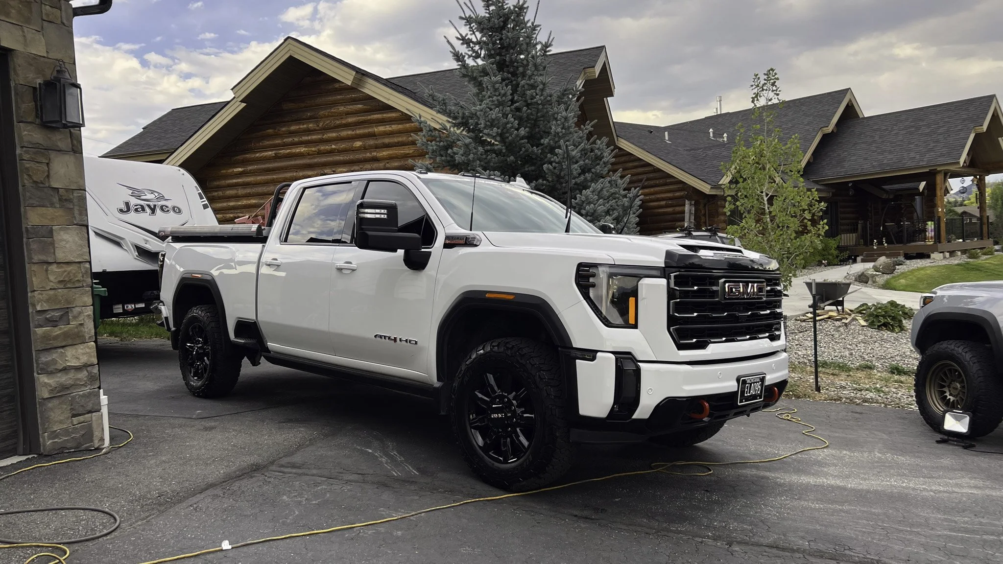 White GMC pickup truck parked outside a house with logs for siding, pull into the driveway, presence of a nearby camper trailer, a tree and landscaping in the background.