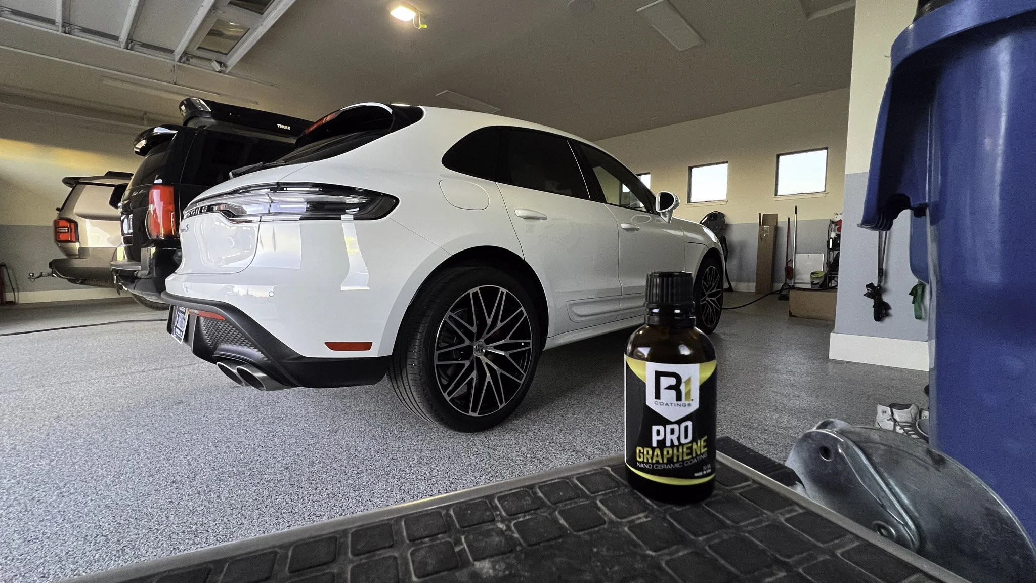 A white luxury SUV parked in a garage with several other vehicles. A bottle of pro graphene nano ceramic coating is in the foreground on a black mat. The garage has gray carpeted floors, windows, and garage tools in the background.