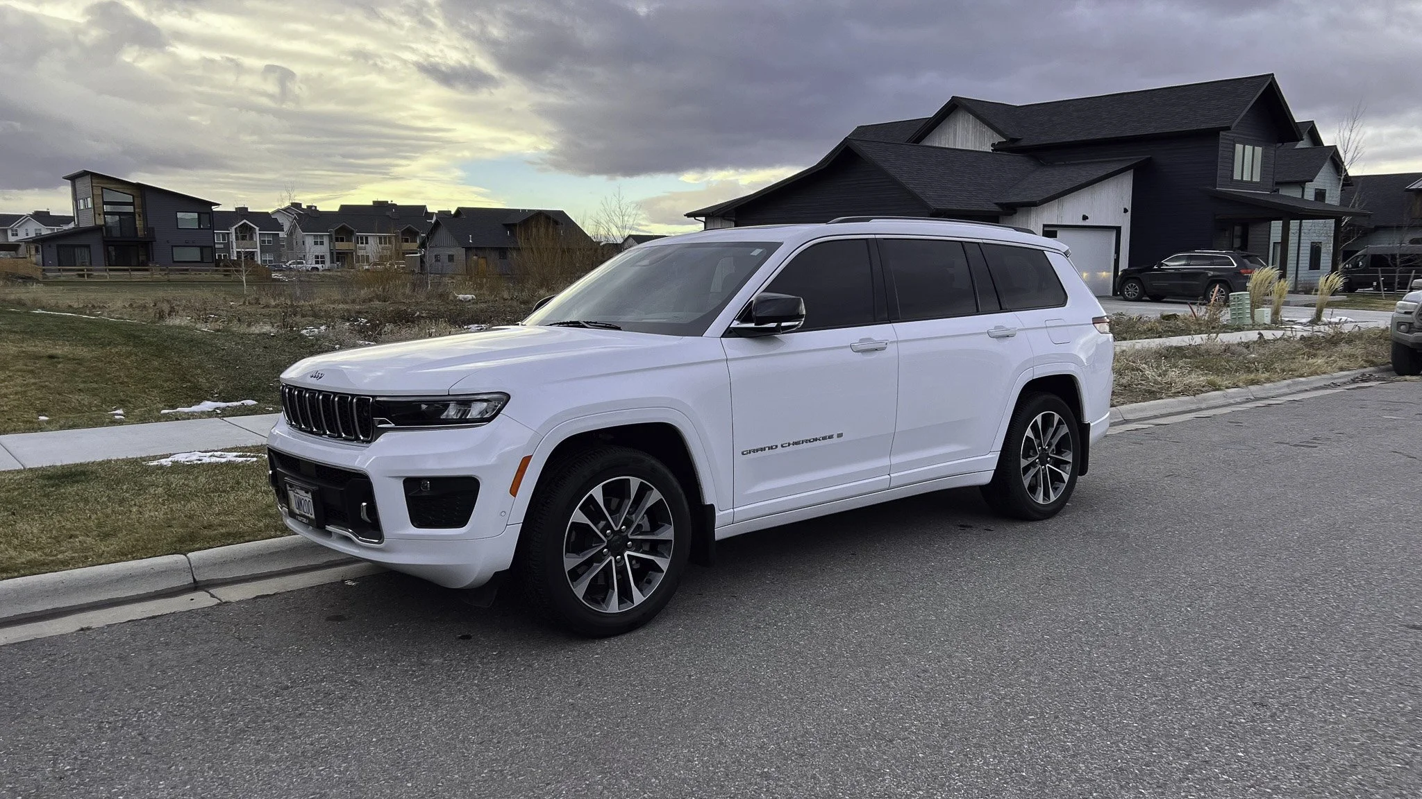 White Jeep Grand Cherokee parked on a residential street with modern houses in the background under a cloudy sky.