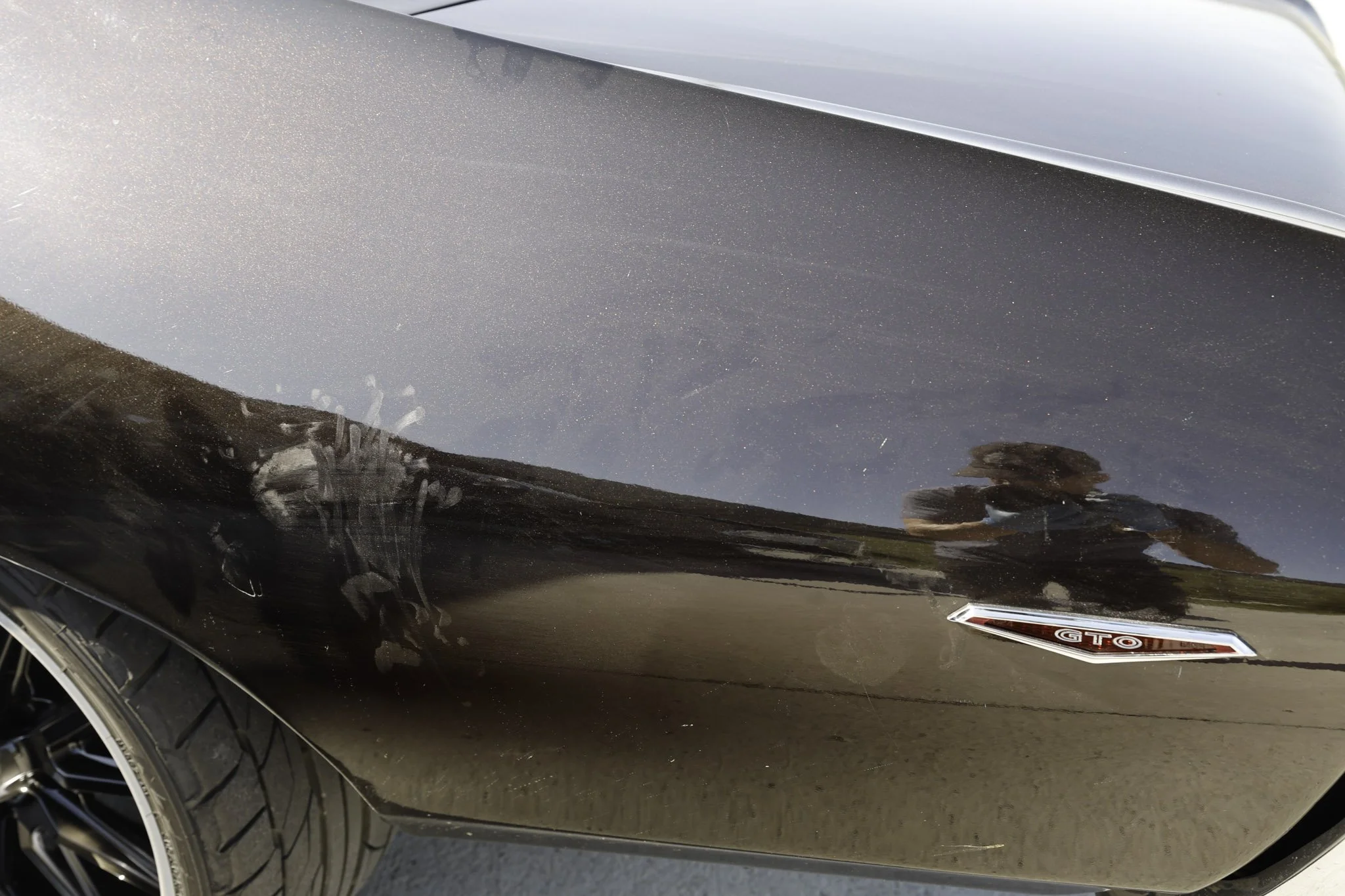 Close-up of a black car's side, reflecting a man taking a photo, with a partial tire visible and a GTO emblem on the side.