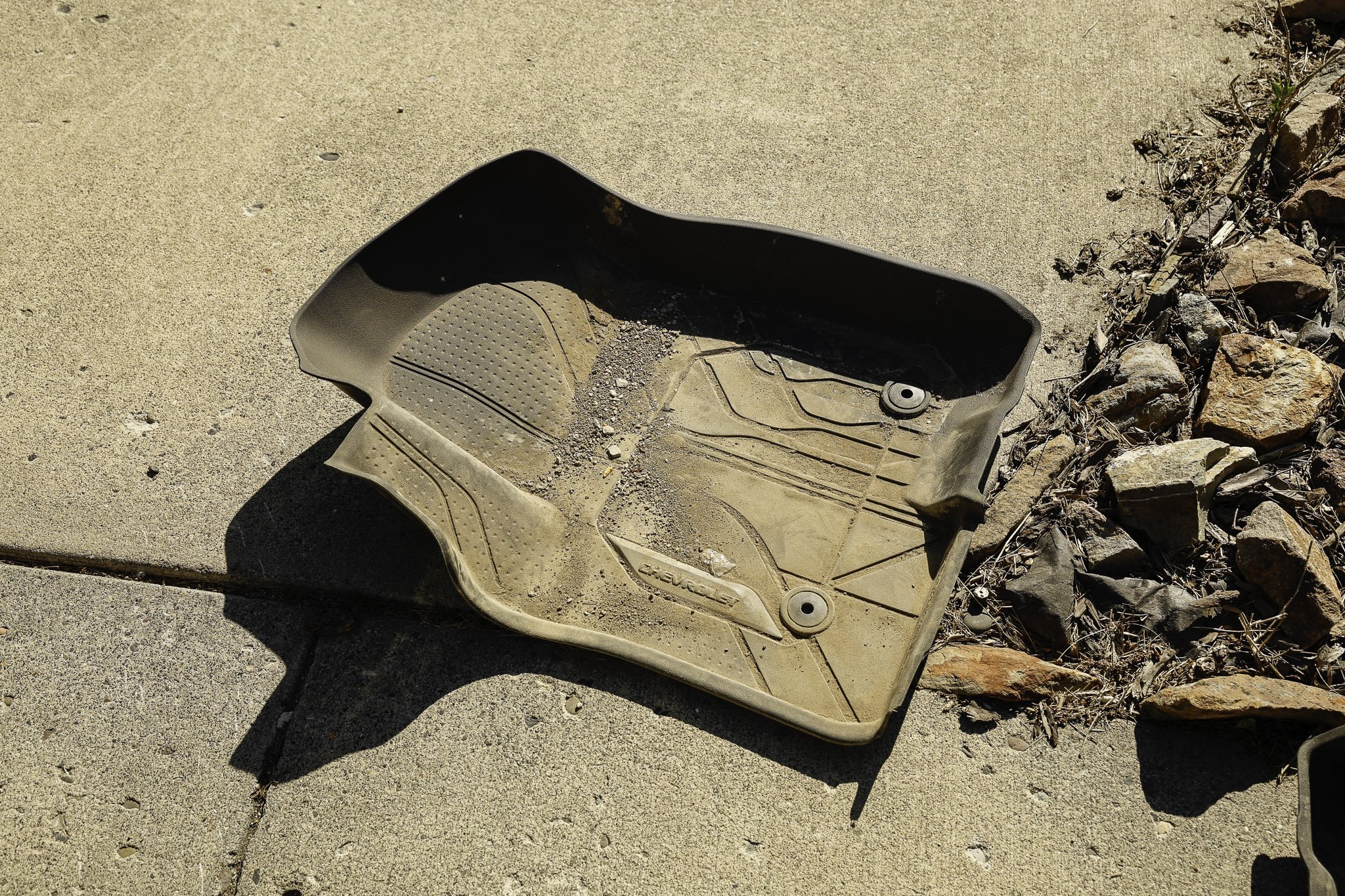 A dusty, dirty car floor mat lying on a concrete sidewalk next to a patch of rocks and small plants.