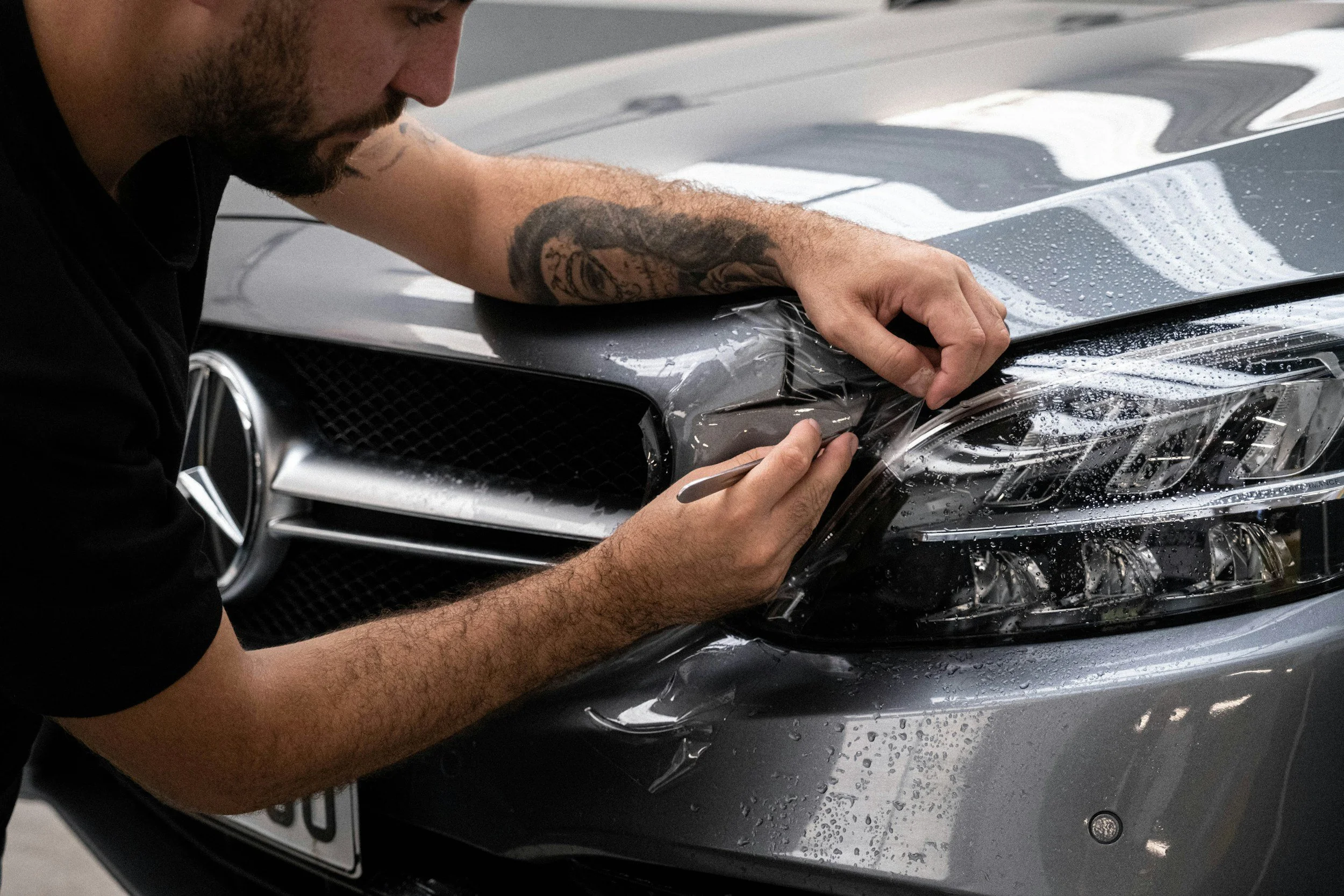 A man applying a protective film or tint to the front of a grey Mercedes-Benz car, focusing on the headlight area.