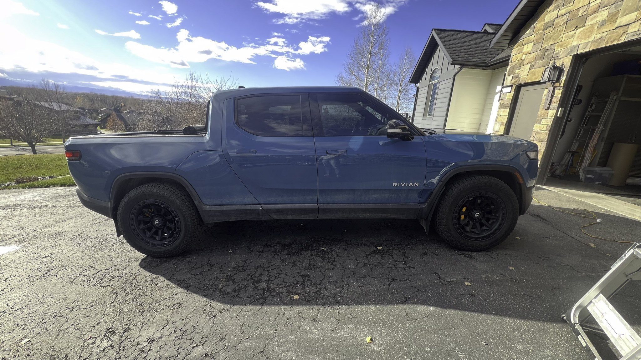 Blue Rivian R1T pickup truck parked in driveway outside a house with stone and siding exterior, under a partly cloudy sky.