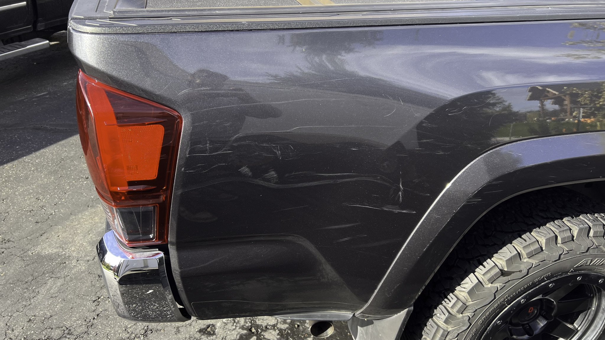 Back right corner of a black pickup truck with scratches on the rear panel, red tail light, chrome bumper, and a tire with an all-terrain tread pattern.