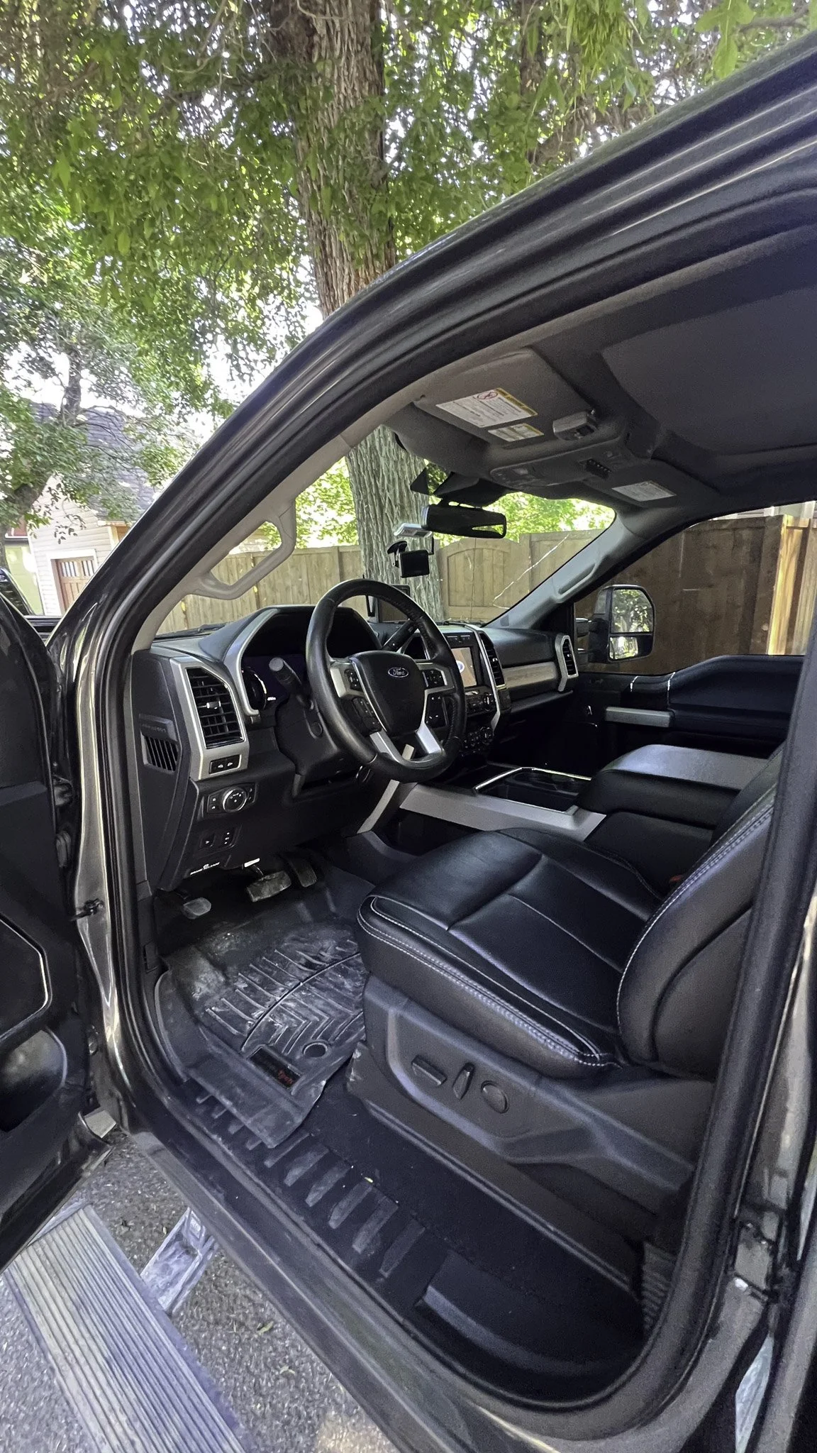 The interior of a black Ford truck, showing the driver's seat, steering wheel, dashboard, and front passenger seat, with trees and a wooden fence visible through the windows.