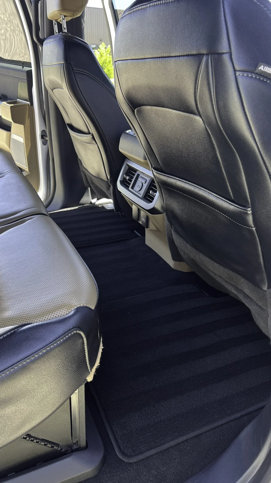 The interior of a vehicle showing the back of front seats, air vents, and black floor mats.
