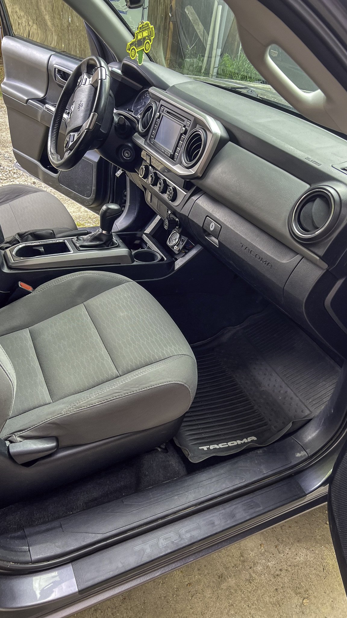 Front interior of a gray Toyota Tacoma pickup truck showing the steering wheel, dashboard, center console, gear shift, and front passenger seat, with a Tacoma-branded floor mat.