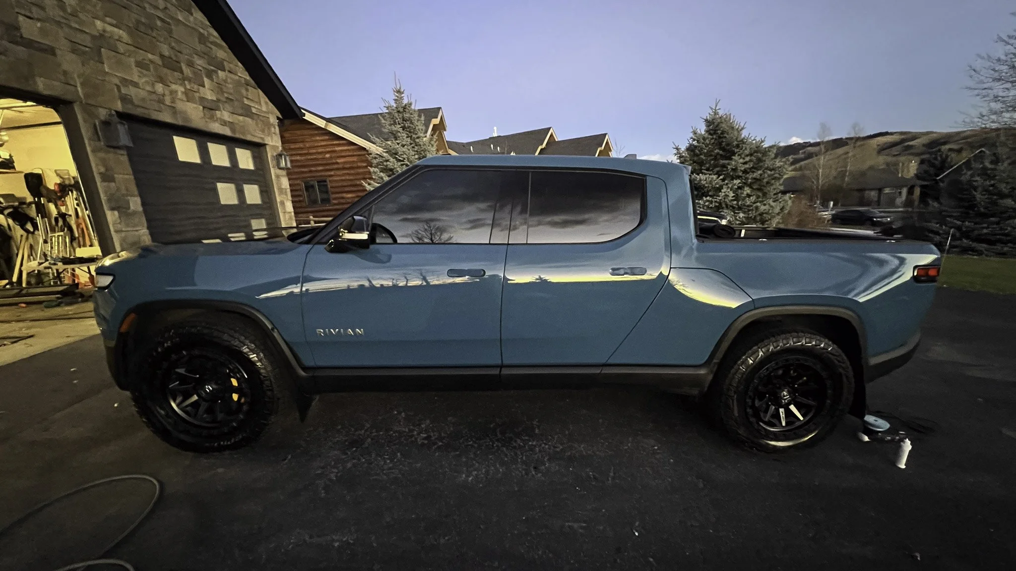 Blue Rivian electric pickup truck parked on a driveway during dusk, with reflective windows showing a cloudy sky, and a garage open to the left with tools inside, on a residential street with trees and houses in the background.