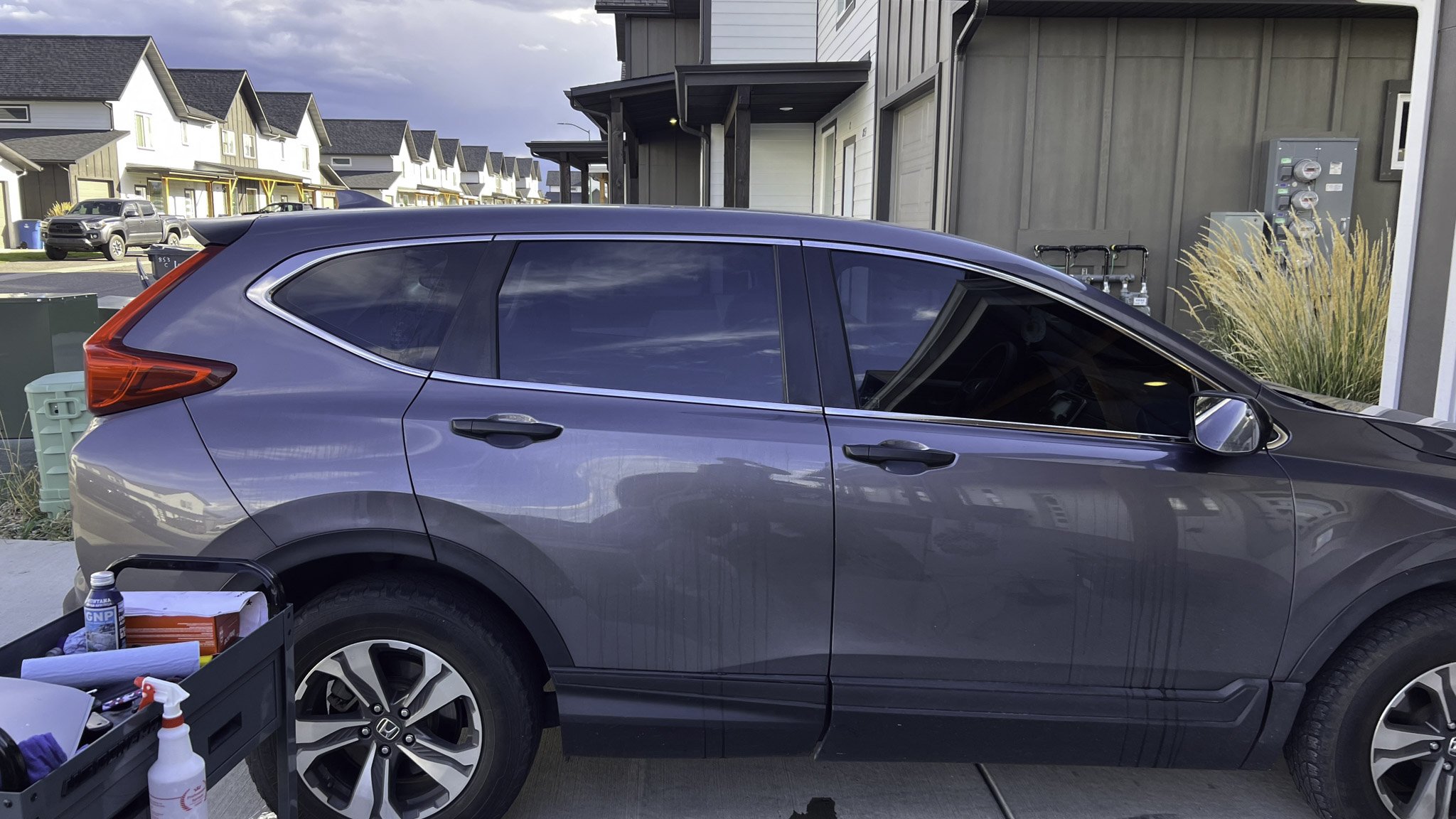 A gray Honda CR-V parked in a residential driveway with houses and a parking lot in the background.