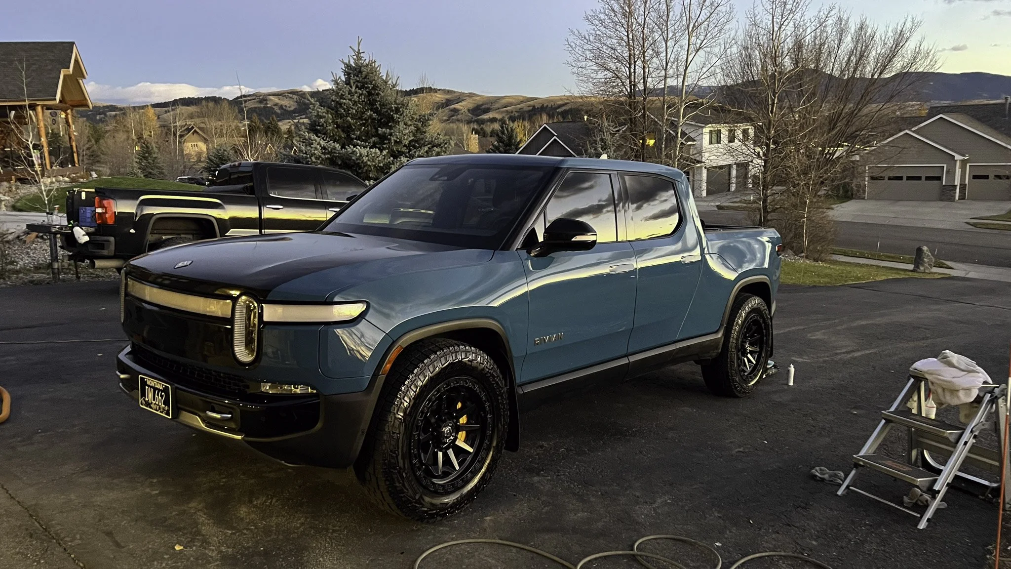 A blue Rivian electric pickup truck parked on a driveway with mountains and houses in the background during sunset.