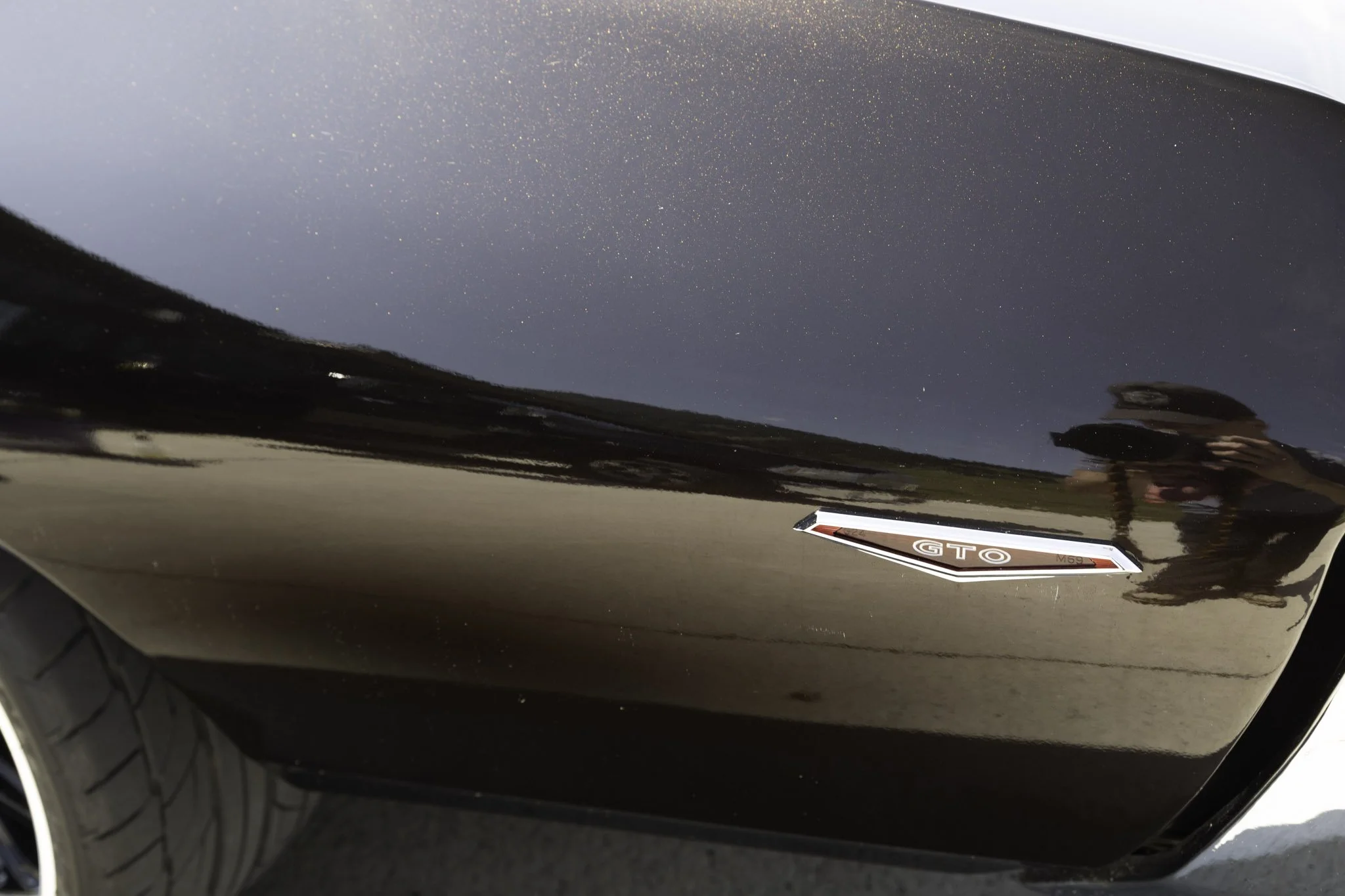 Close-up of the side of a dark-colored GMC GTO car, showcasing the GTO badge and a reflection of the person taking the photo.