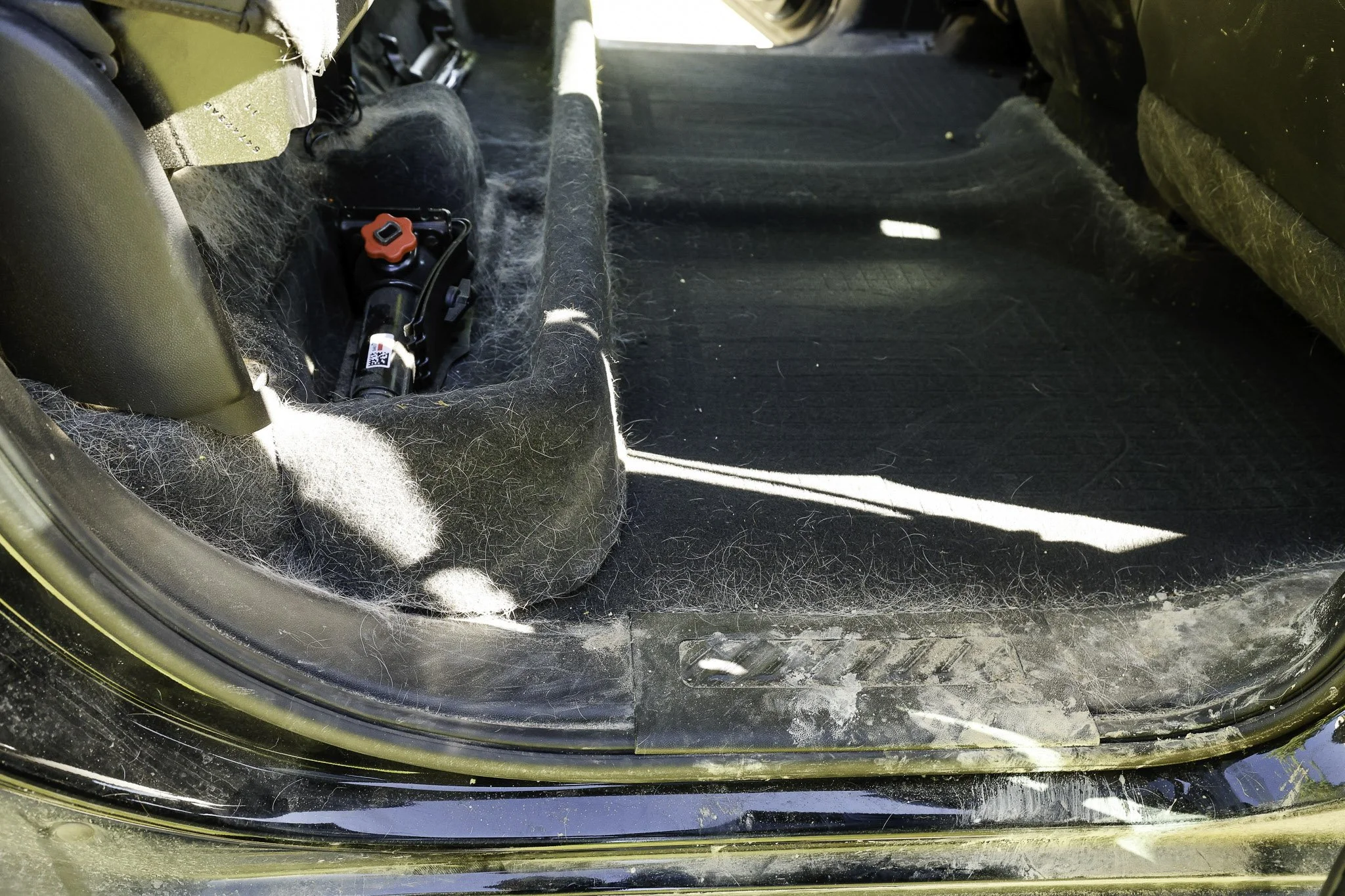 The interior of a vehicle showing the empty back seat area with a black floor mat, some dirt, and a tool or device with red and black parts on the left side.