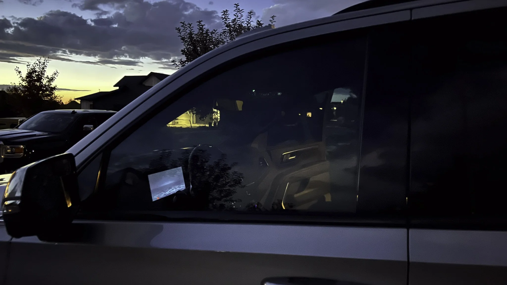A dashboard and steering wheel visible inside a parked vehicle during dusk, with other cars and houses in the background, and a cloudy sky at sunset.