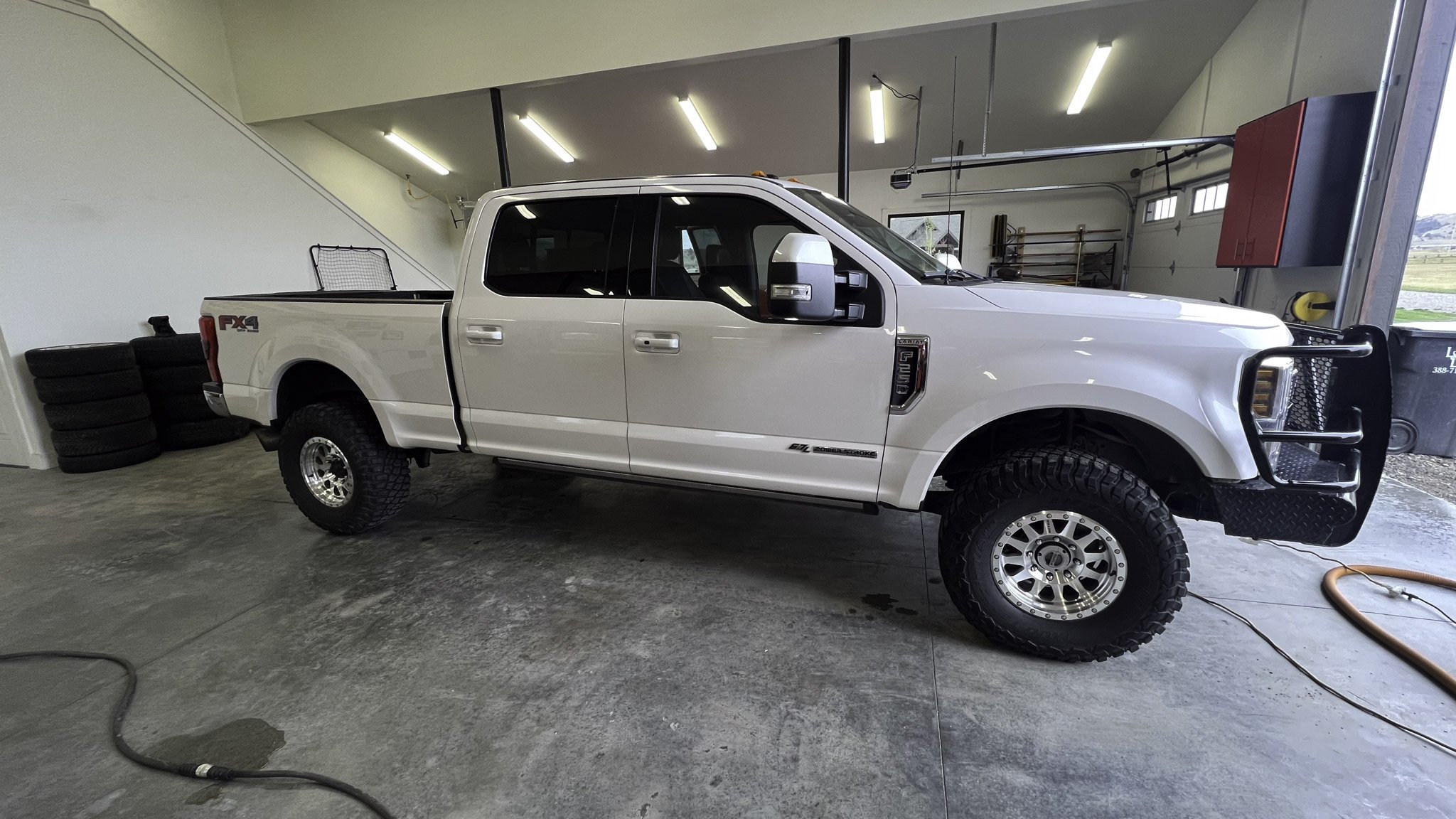 White four-door pickup truck with off-road tires, parked inside a garage with a concrete floor, stacked tires, and various tools and shelves in the background.