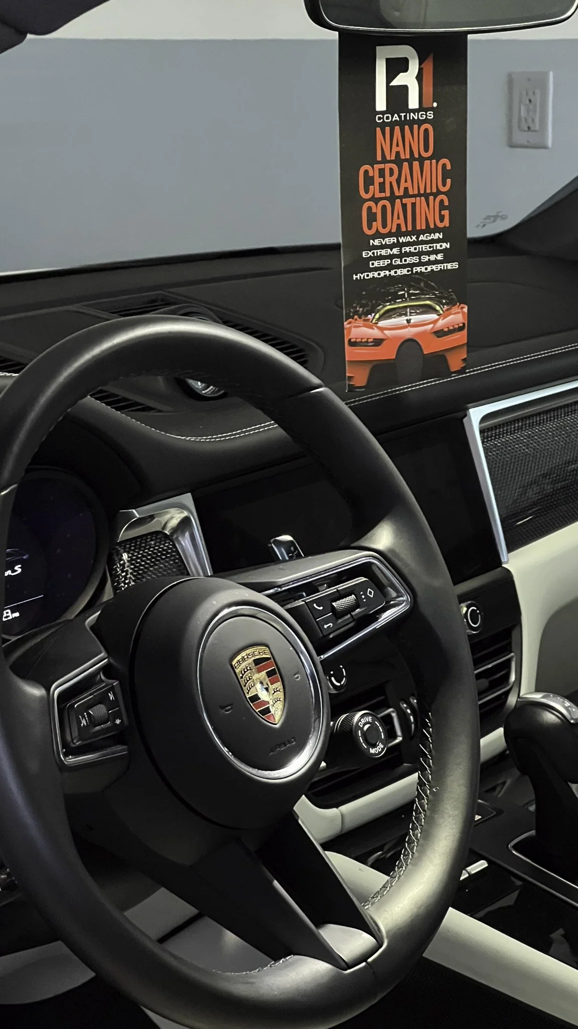 The interior of a Porsche vehicle, showing the steering wheel with Porsche logo and controls, and a hanging car care product labeled 'R1 Coatings Nano Ceramic Coating' with a red sports car image.