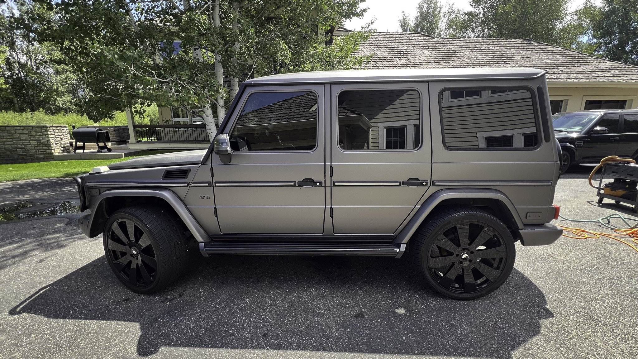 A gray Mercedes Benz G-Class SUV parked outside on a paved driveway with a house and trees in the background.