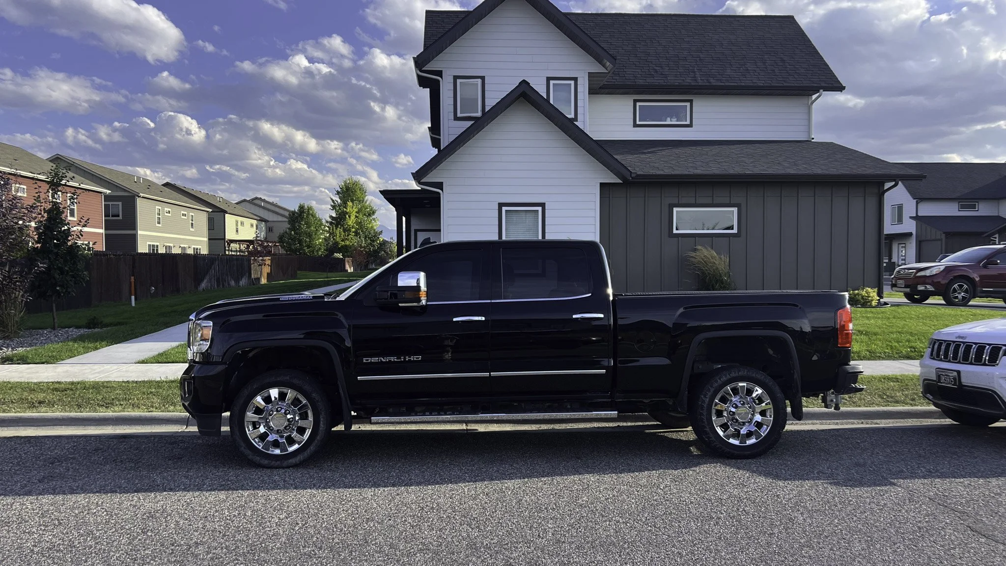 A black GMC Sierra Denali HD pickup truck parked on a residential street in front of a modern house with white and gray siding. Other cars and houses are visible in the background under a partly cloudy sky.