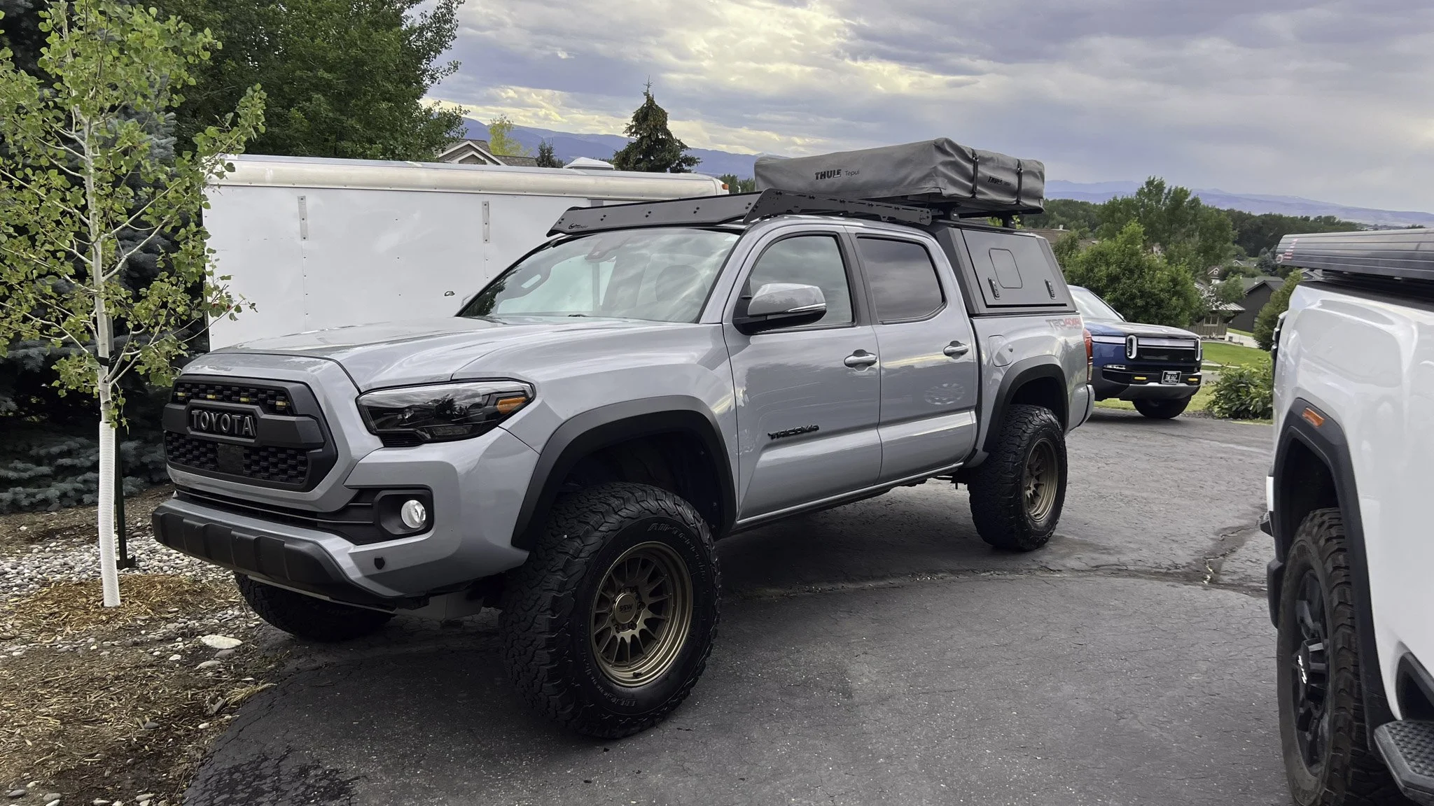 Gray Toyota pickup truck with off-road tires, mounted roof storage, and camper shell parked on a driveway with a white trailer and trees in the background.