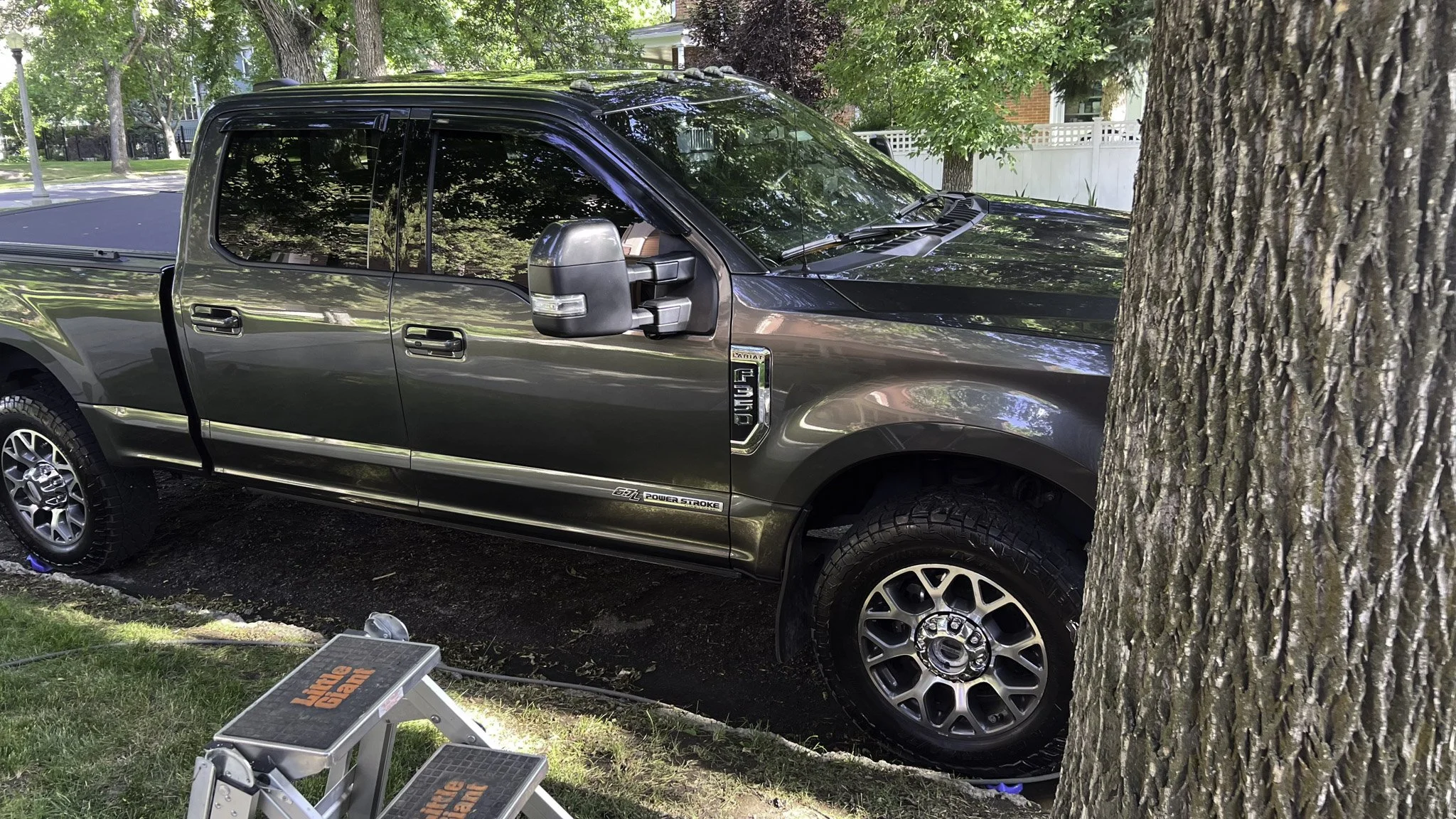 A dark gray Ford F-250 pickup truck parked on a grassy patch next to a tree. The front end of the truck is partially hidden behind the tree. The truck's windows are reflective, showing the surrounding trees and sky. There is a small step stool with o