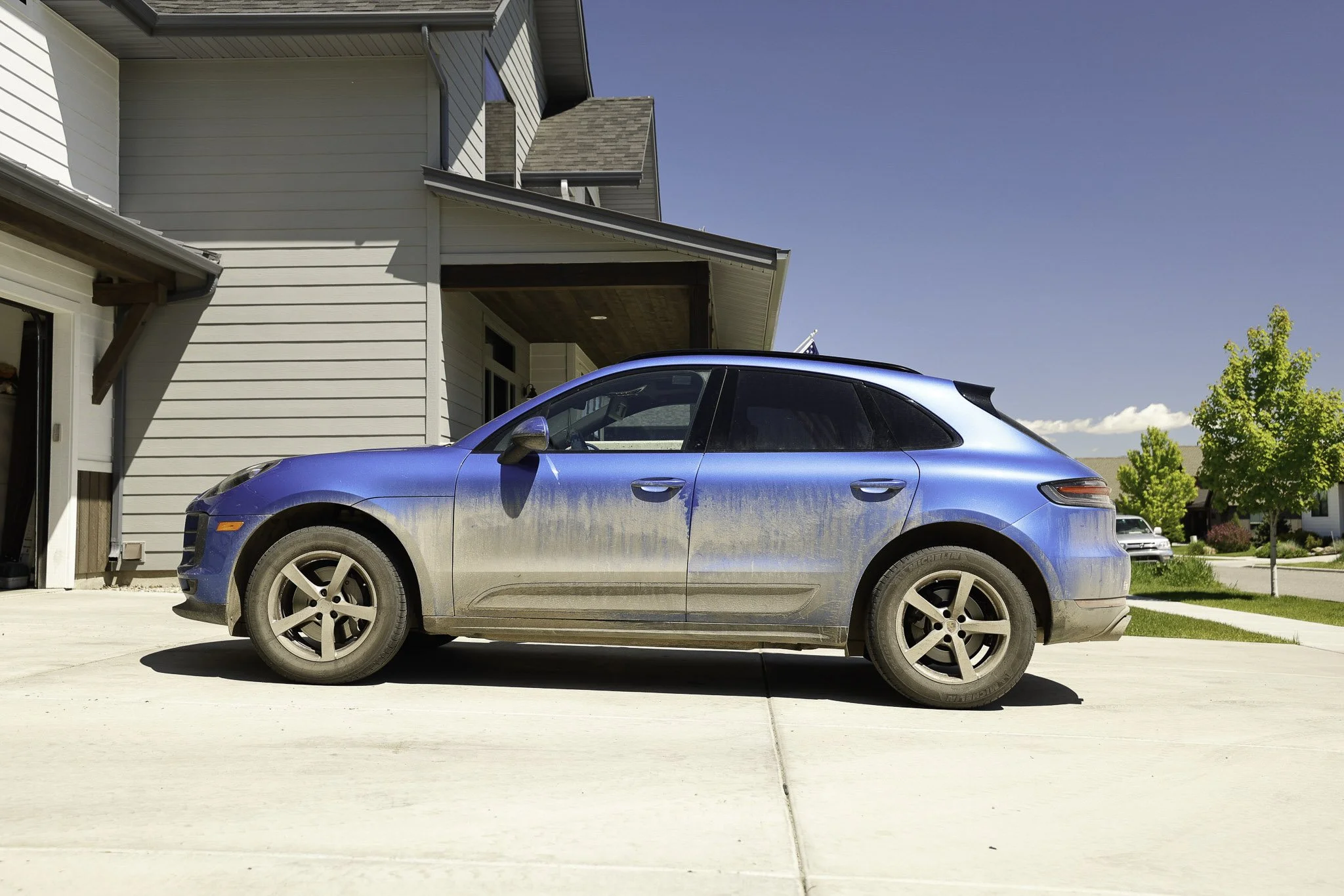 A blue SUV parked in a driveway in front of a modern house with gray siding and a sloped roof. The SUV is dirty with dust and mud on the sides, wheel areas, and lower body. Trees and a clear blue sky are visible in the background.
