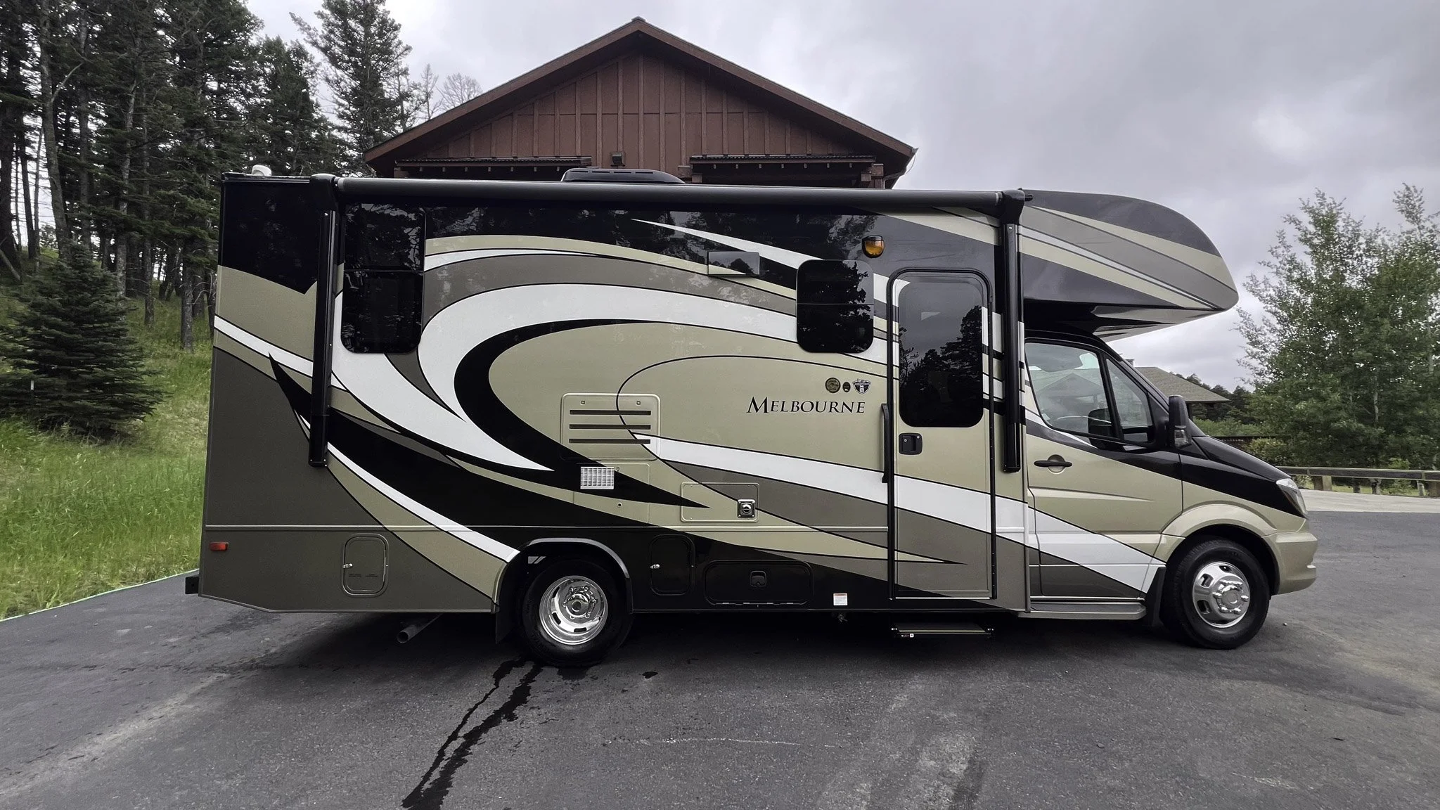 A beige and black motorhome RV parked on a paved surface with green grass, trees, and a red wooden building in the background under cloudy skies.