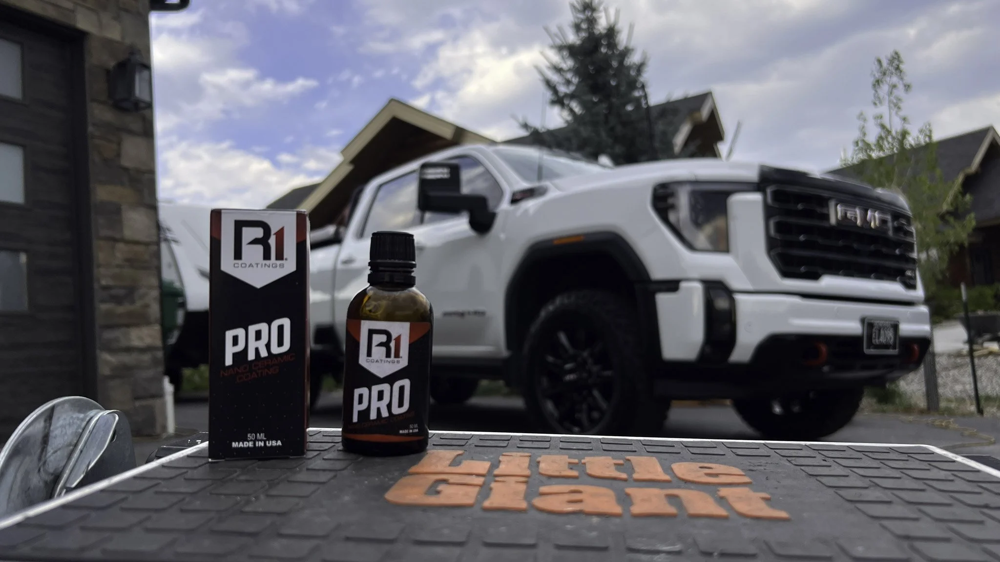 A white GMC truck parked outside a house with a garage. In the foreground, there is a bottle of R1 PRO coating and its box, placed on a textured mat that says 'Little Giant'.