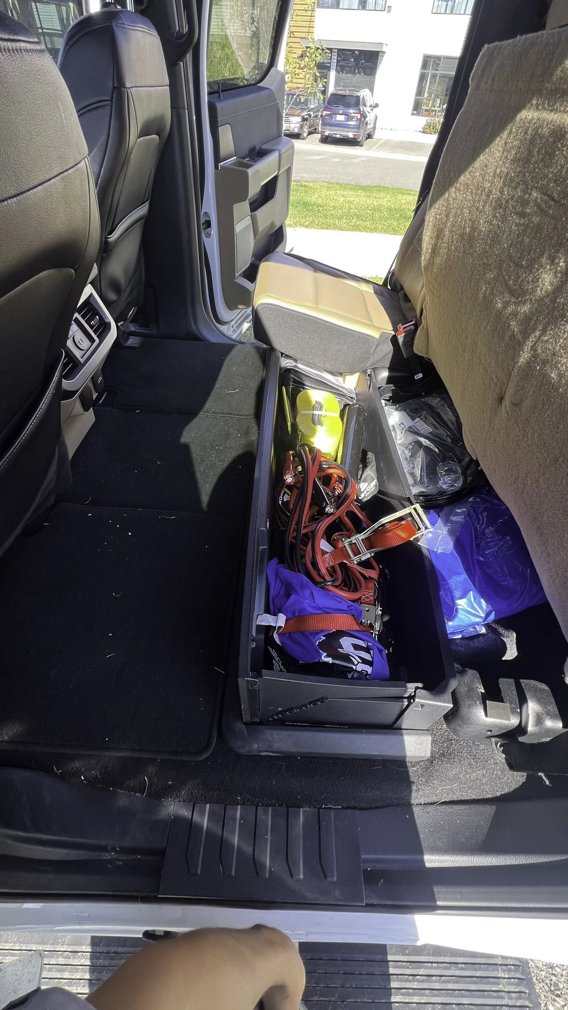 View inside a vehicle showing a storage bin with red and black cables, a blue bag, yellow objects, and some plastic bags on the backseat.