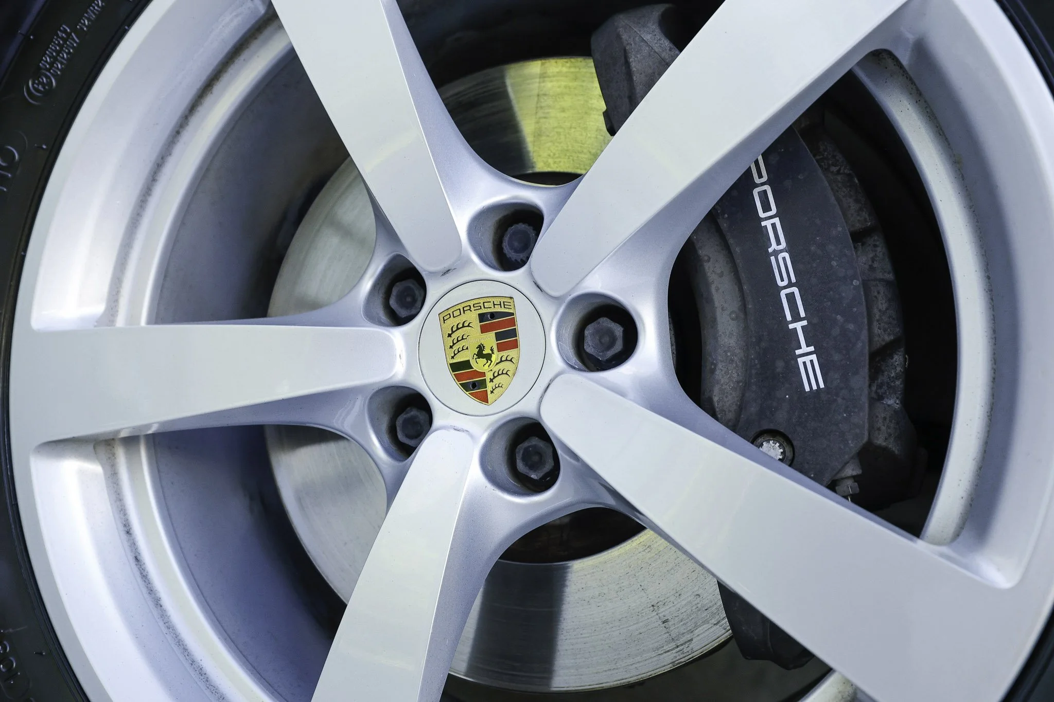 Close-up of a Porsche wheel with a silver rim and the Porsche emblem in the center, showing brake discs and calipers behind the spokes.