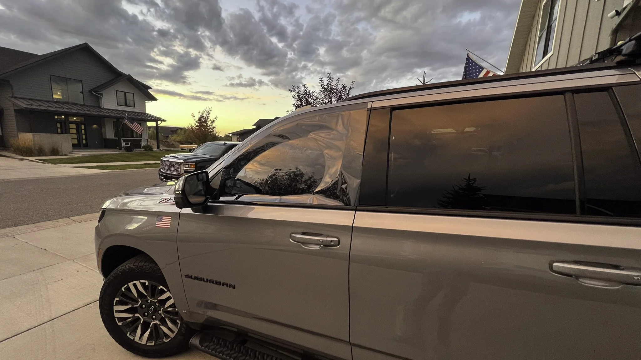 A silver Subaru SUV parked on a driveway during sunset, with an American flag on the front fender and a town house with an American flag and trees in the background.
