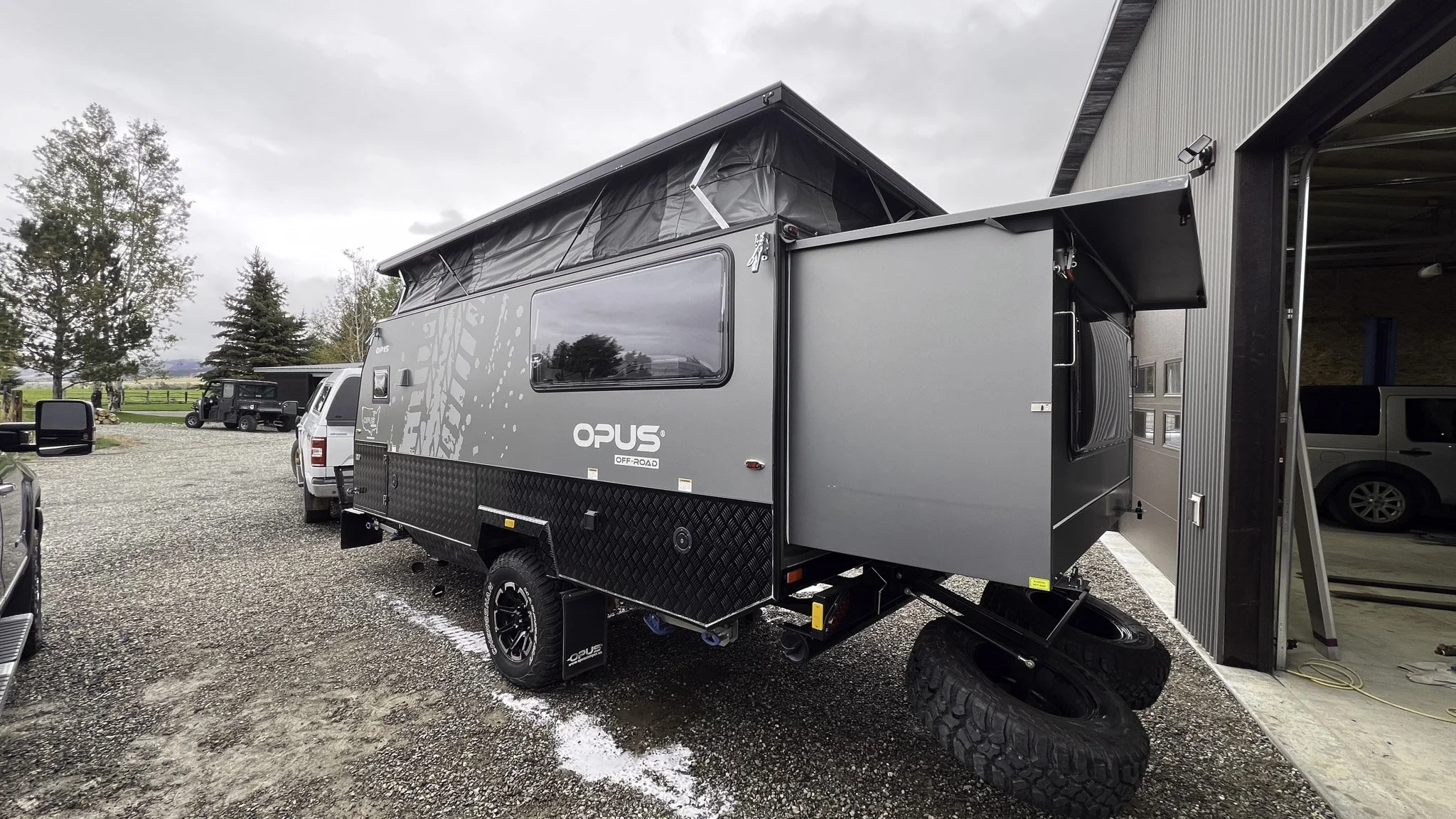A gray off-road camper trailer with an extended slide-out section parked outside a garage, with tires and a pop-up roof tent visible, and several other vehicles in the background on a gravel lot.