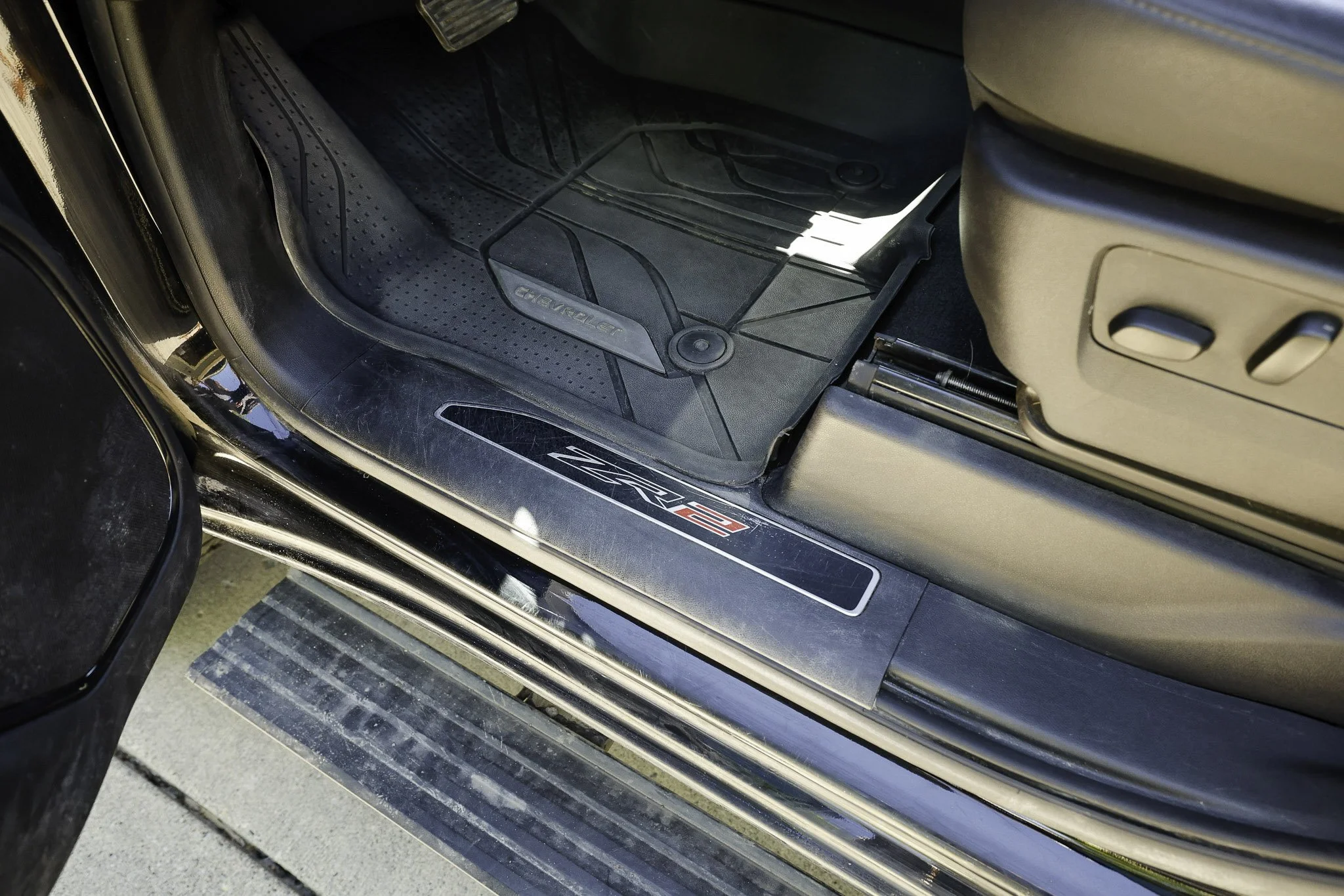 Close-up of the interior door sill and floor area of a black Chevrolet vehicle, showing a rubber floor mat and part of the seat adjustment controls.
