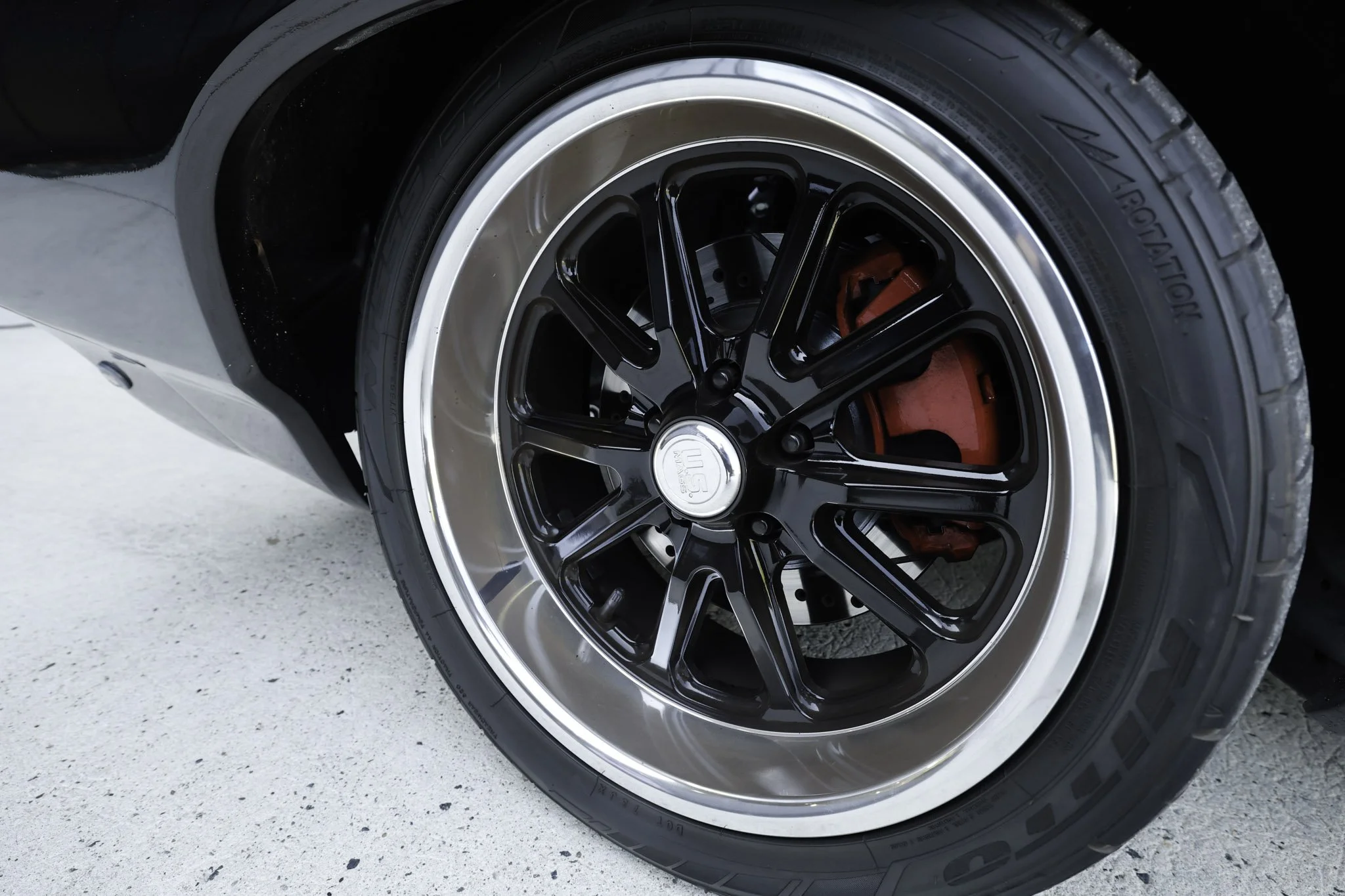 Close-up of a car wheel with a black alloy rim, red brake caliper, and a tire on a light-colored concrete surface.