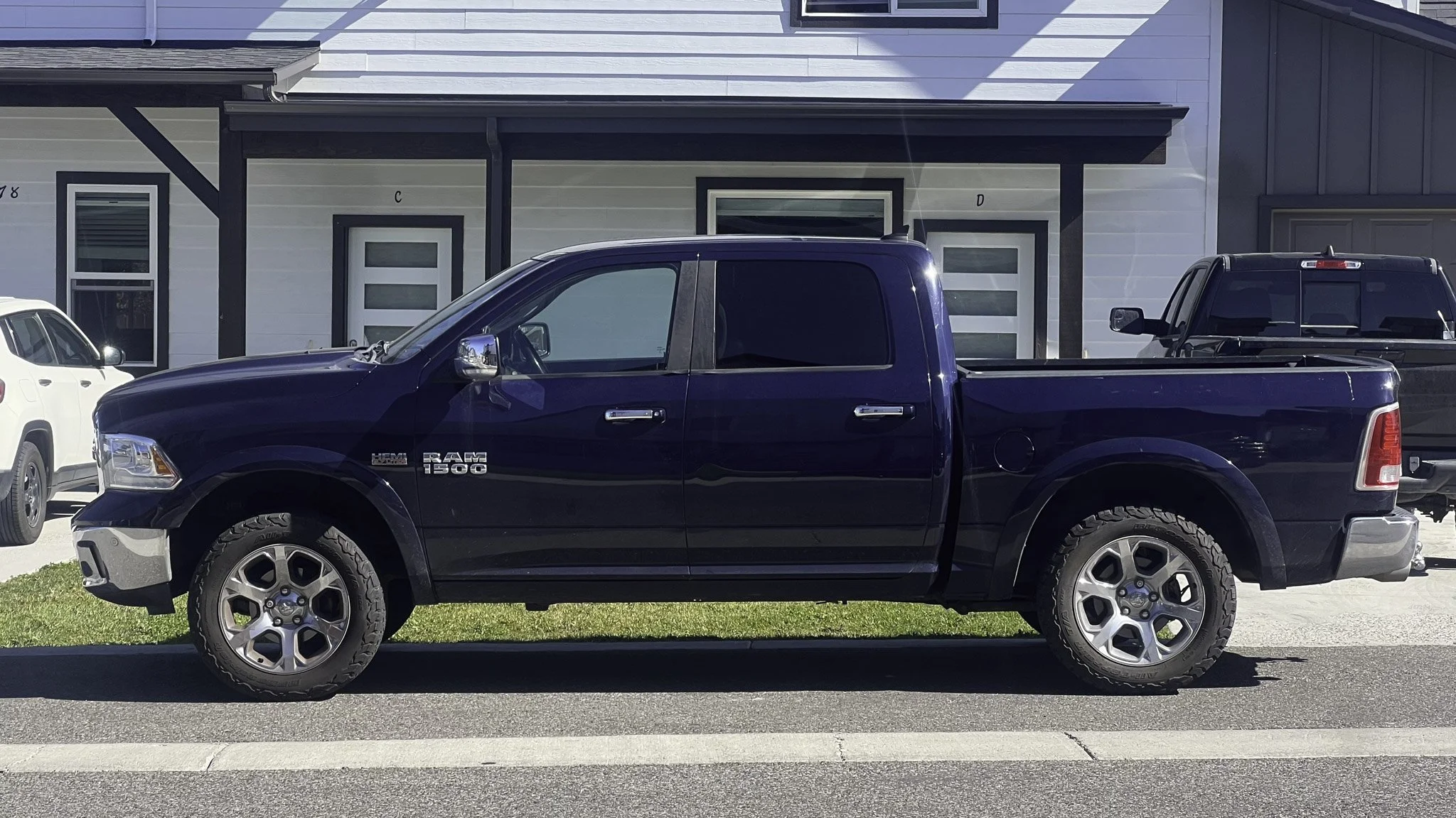 A black RAM 1500 pickup truck parked on the street in front of a modern two-story house with white and dark gray exterior panels. Other vehicles, including a white SUV and a black truck, are visible parked nearby.