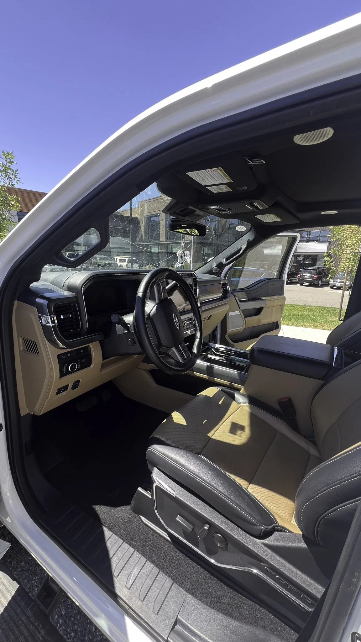 Interior of a modern pickup truck with a black steering wheel, digital dashboard, beige and black seats, and a center console, parked outdoors on a sunny day.