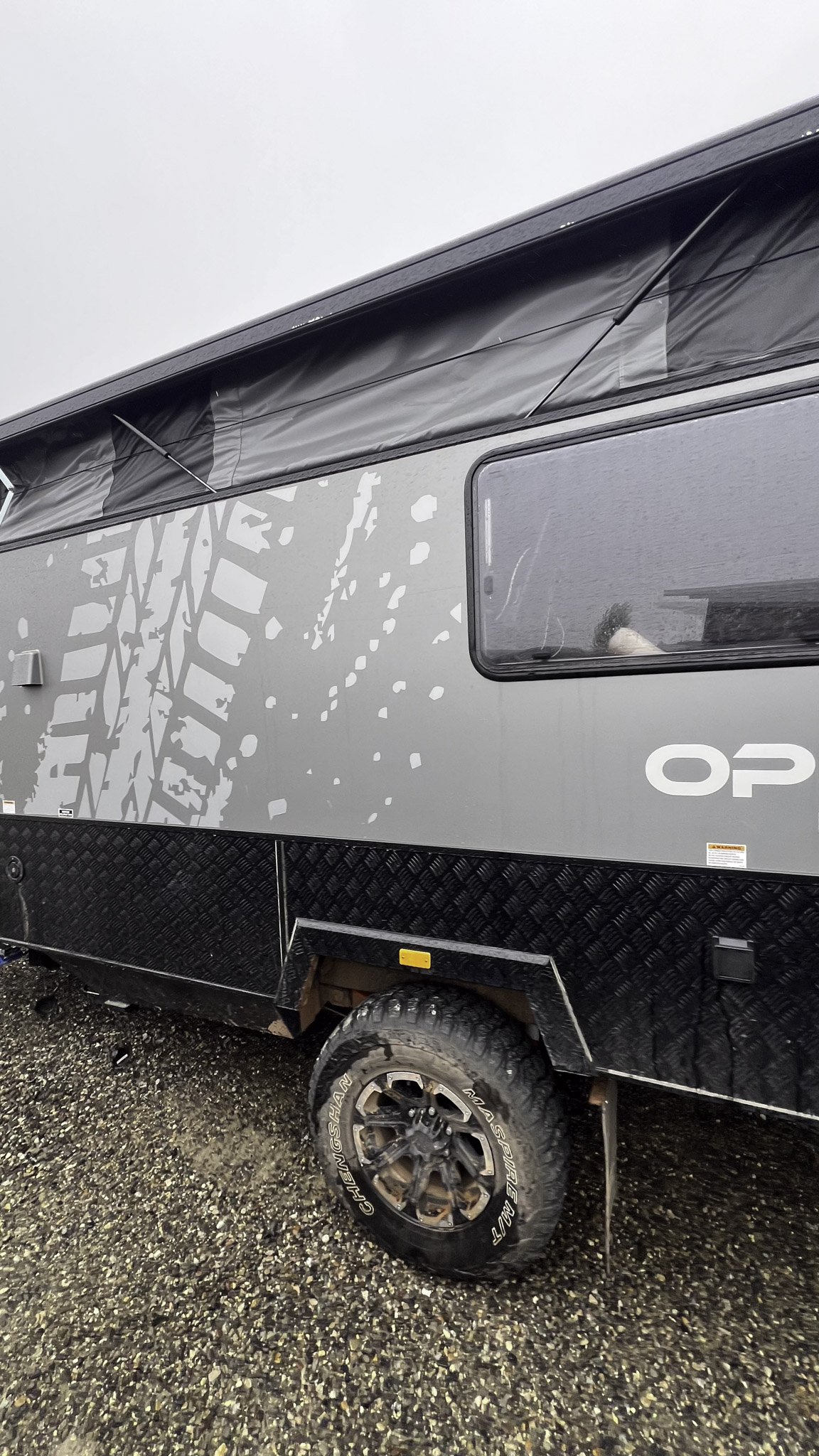 Close-up of a grey and black off-road camper trailer with rugged tires on a gravel surface, and a small window on its side.