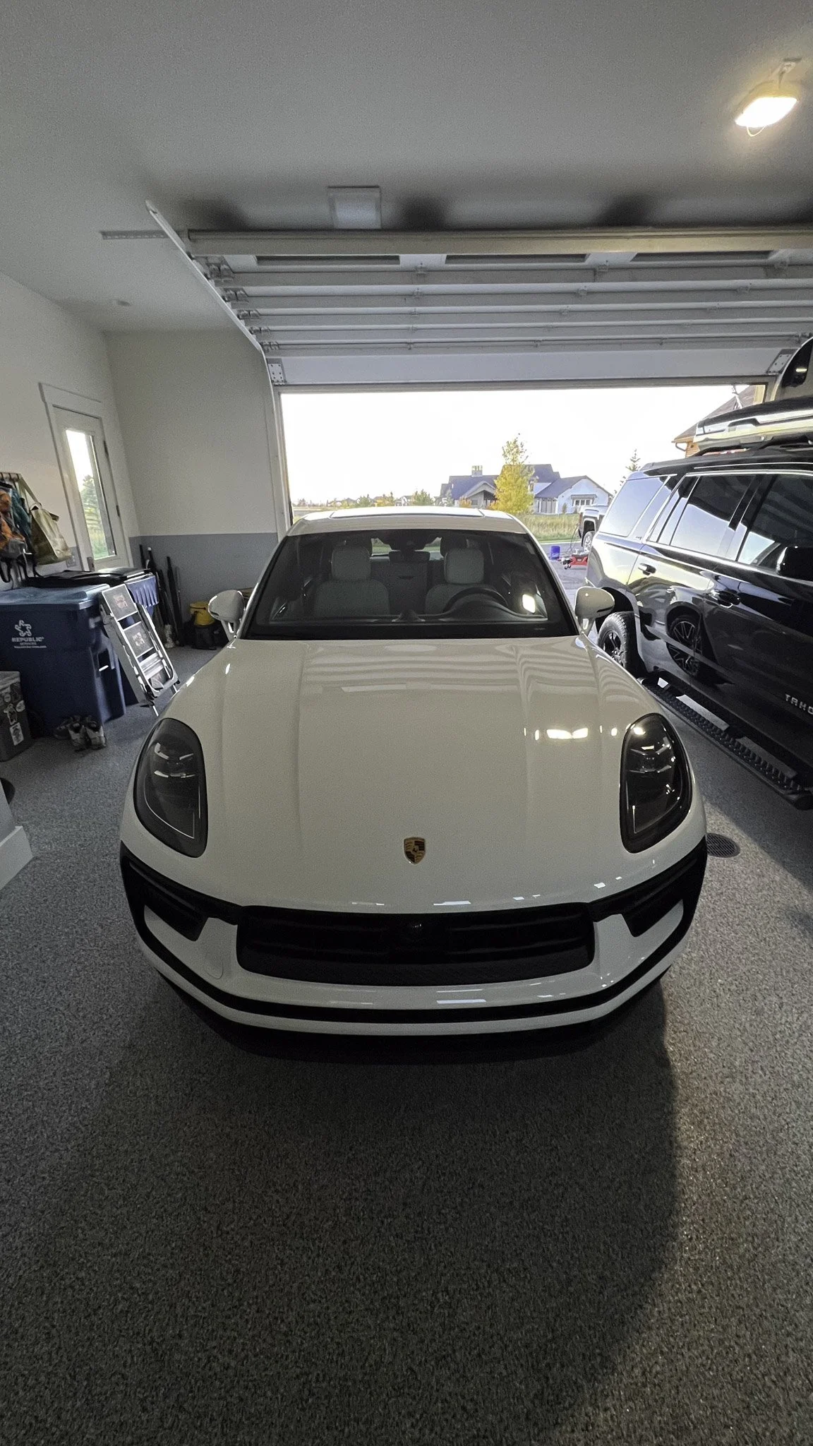 Front view of a white Porsche sports car parked inside a garage with an open roll-up door, showing a residential neighborhood outside.