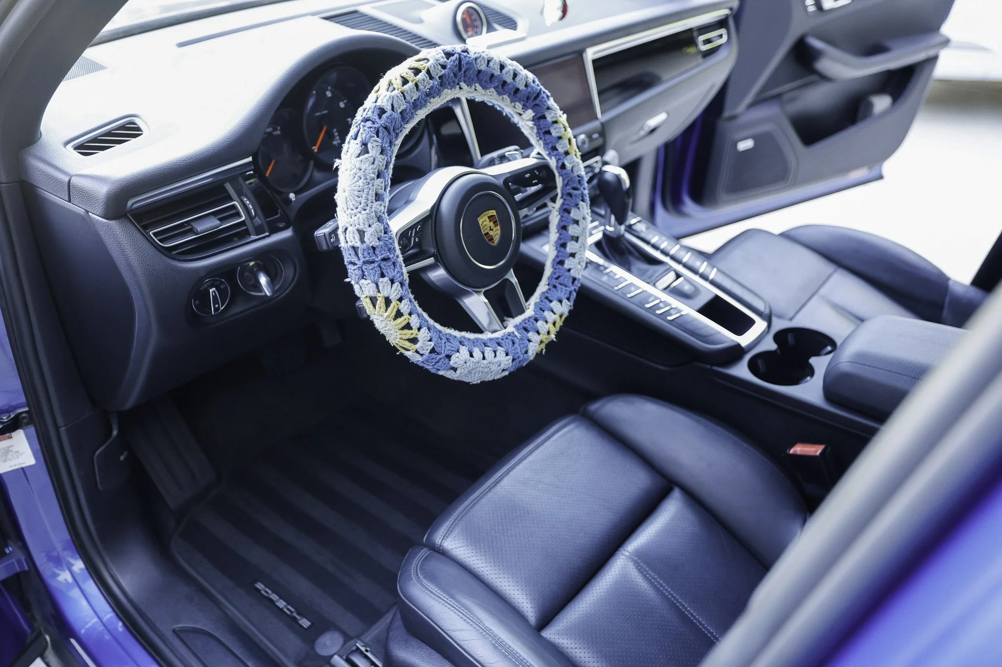Interior of a Porsche vehicle with a crocheted steering wheel cover in blue, white, and yellow. The dashboard, gear shift, and black leather seat are visible.