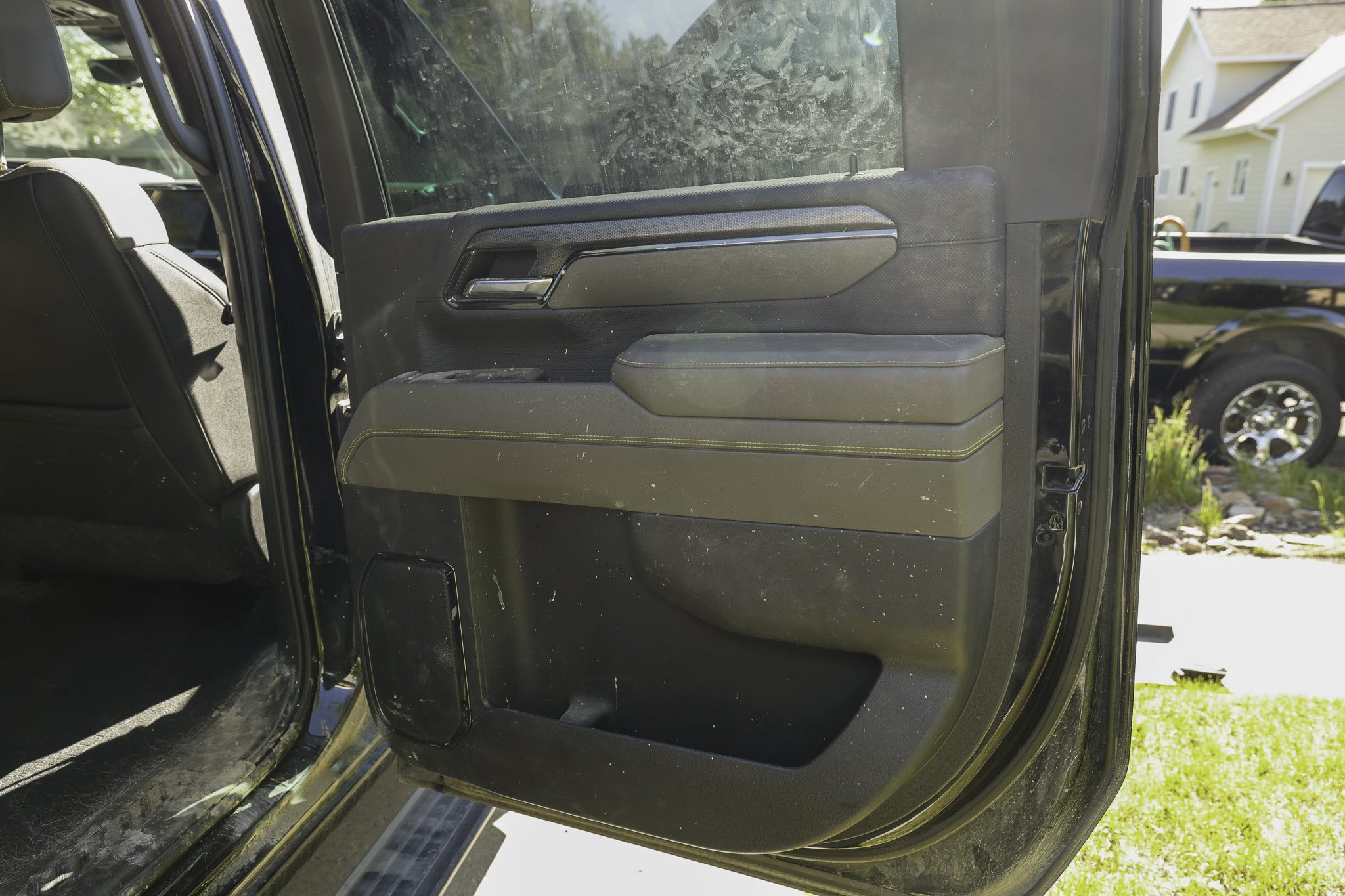 Close-up of a black vehicle's interior driver side door panel with visible handle, armrest, and storage compartment, next to a suburban neighborhood with houses and a black pickup truck outside.