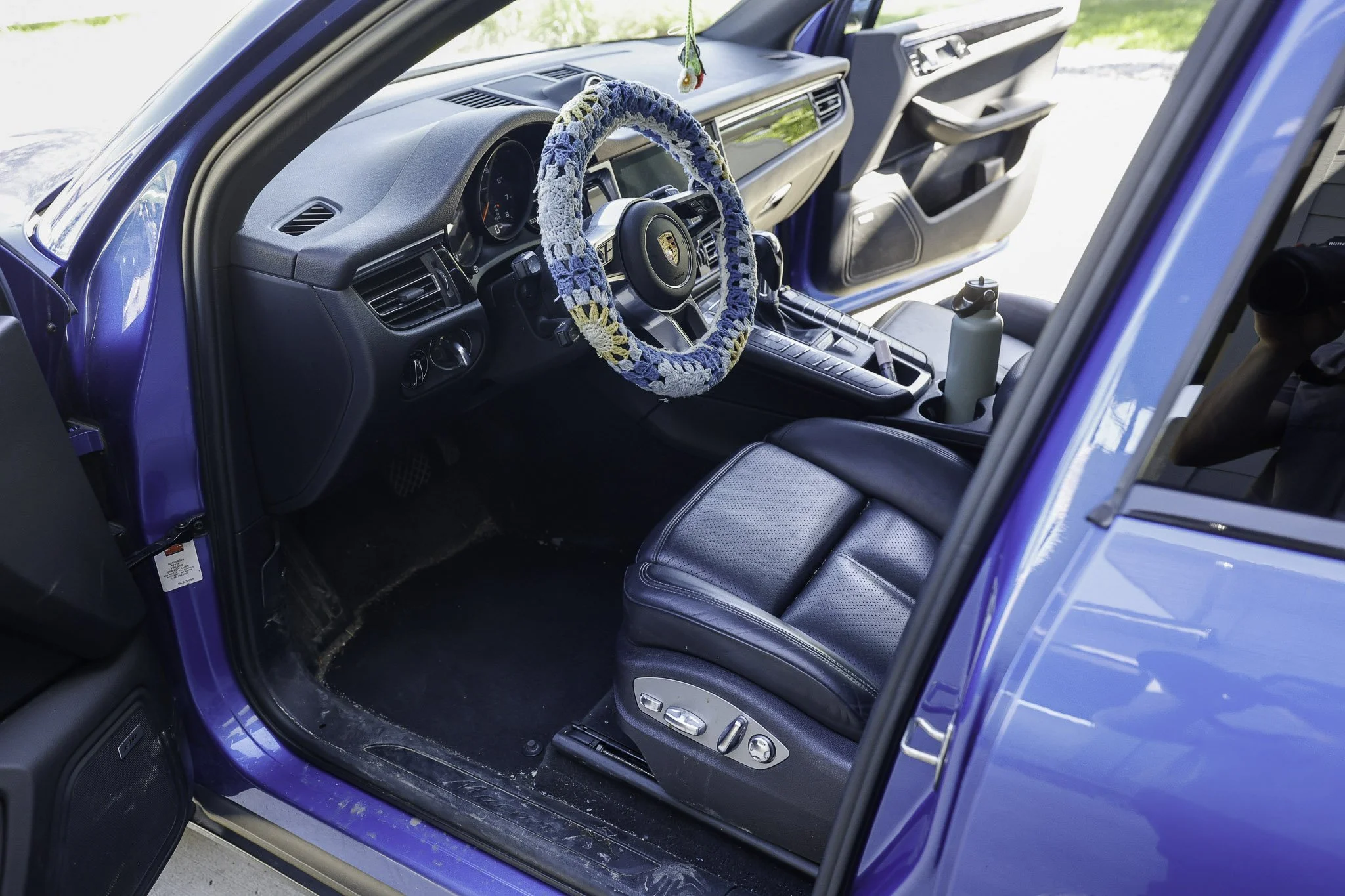Interior view of a blue car showing the driver's seat, steering wheel with a blue and white cover, and the dashboard. There is a water bottle in the cup holder.