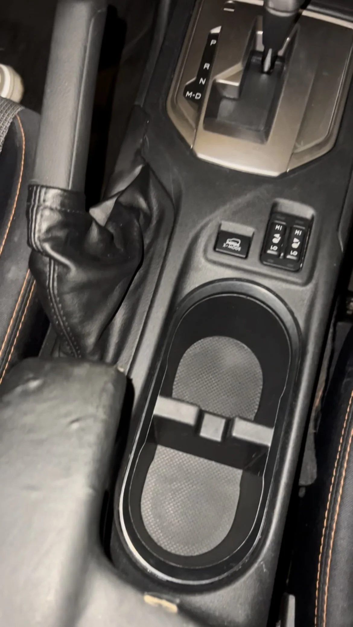 Center console of a vehicle with gear shift, cup holder, control buttons, and part of a seat with orange stitching.