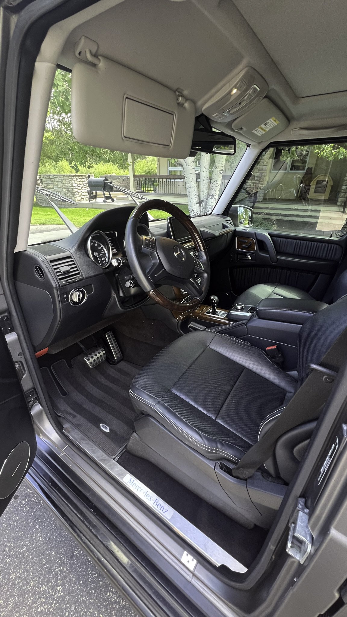 Interior of a Mercedes-Benz vehicle showing the driver's seat, steering wheel, dashboard, gear shifter, and front passenger seat, with a view of the outside greenery through the windows.