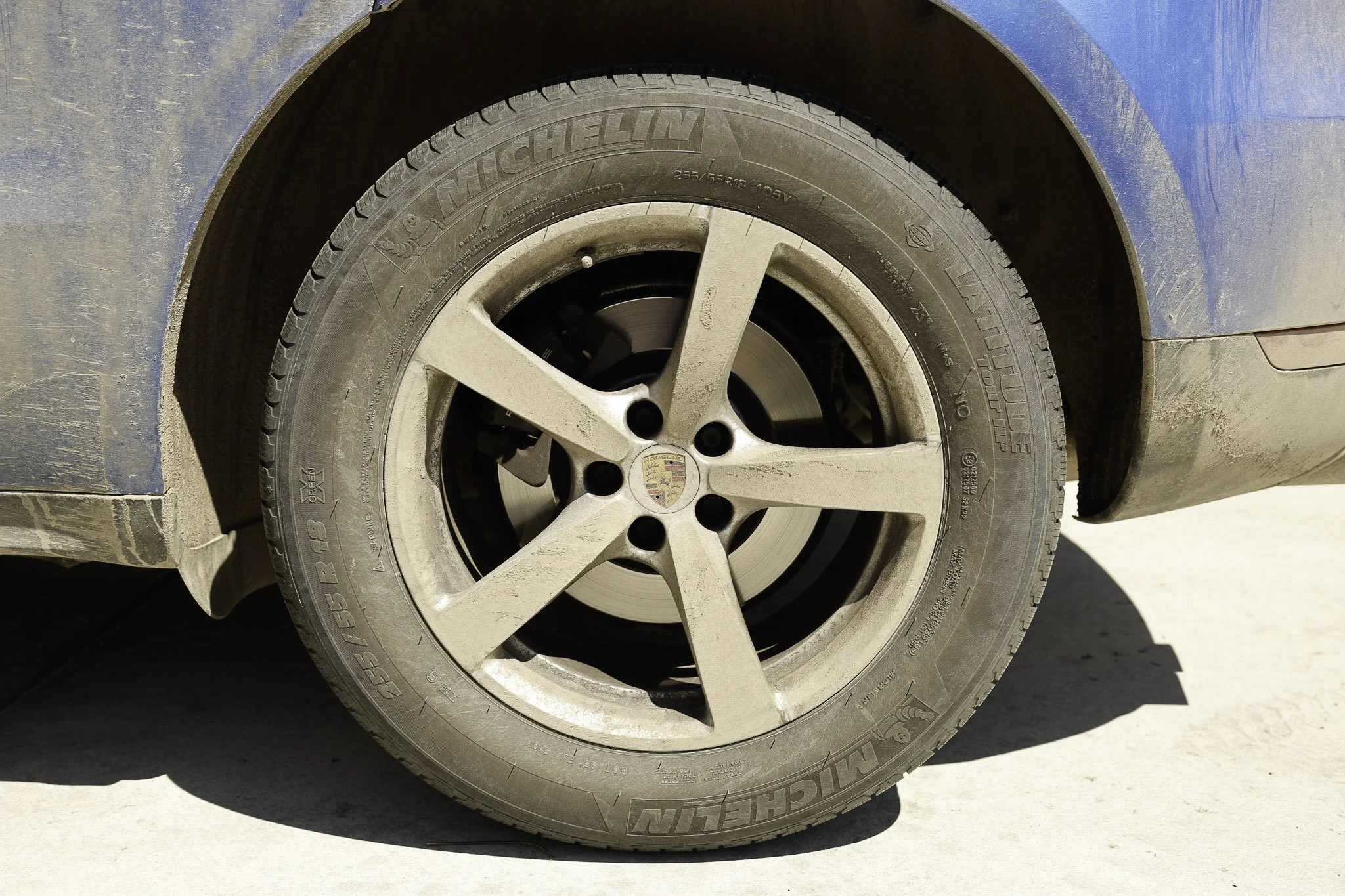 Close-up of a dusty car tire and alloy wheel on a blue vehicle, with the Porsche emblem in the center, parked on a concrete surface, casting a shadow.
